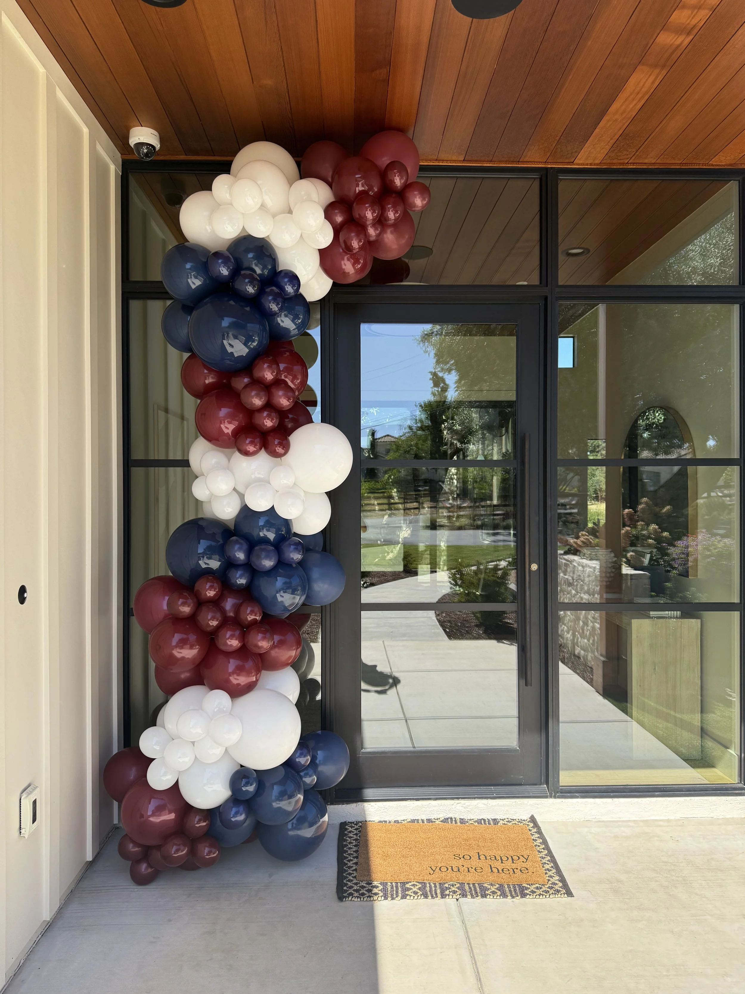 Decorative balloon arch in red, white, and blue colors, placed beside a glass door with a welcome mat reading "so happy you're here." on the porch of a house.
