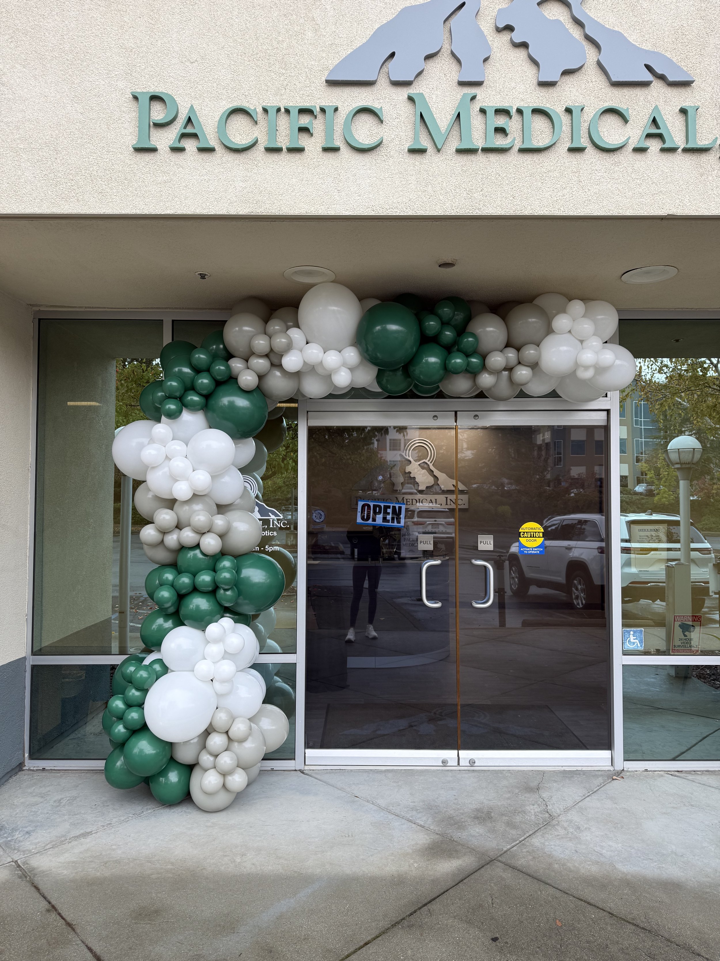 Entrance of Pacific Medical clinic decorated with green, white, and beige balloons in an arch around the door, with a glass door that has an 'Open' sign.