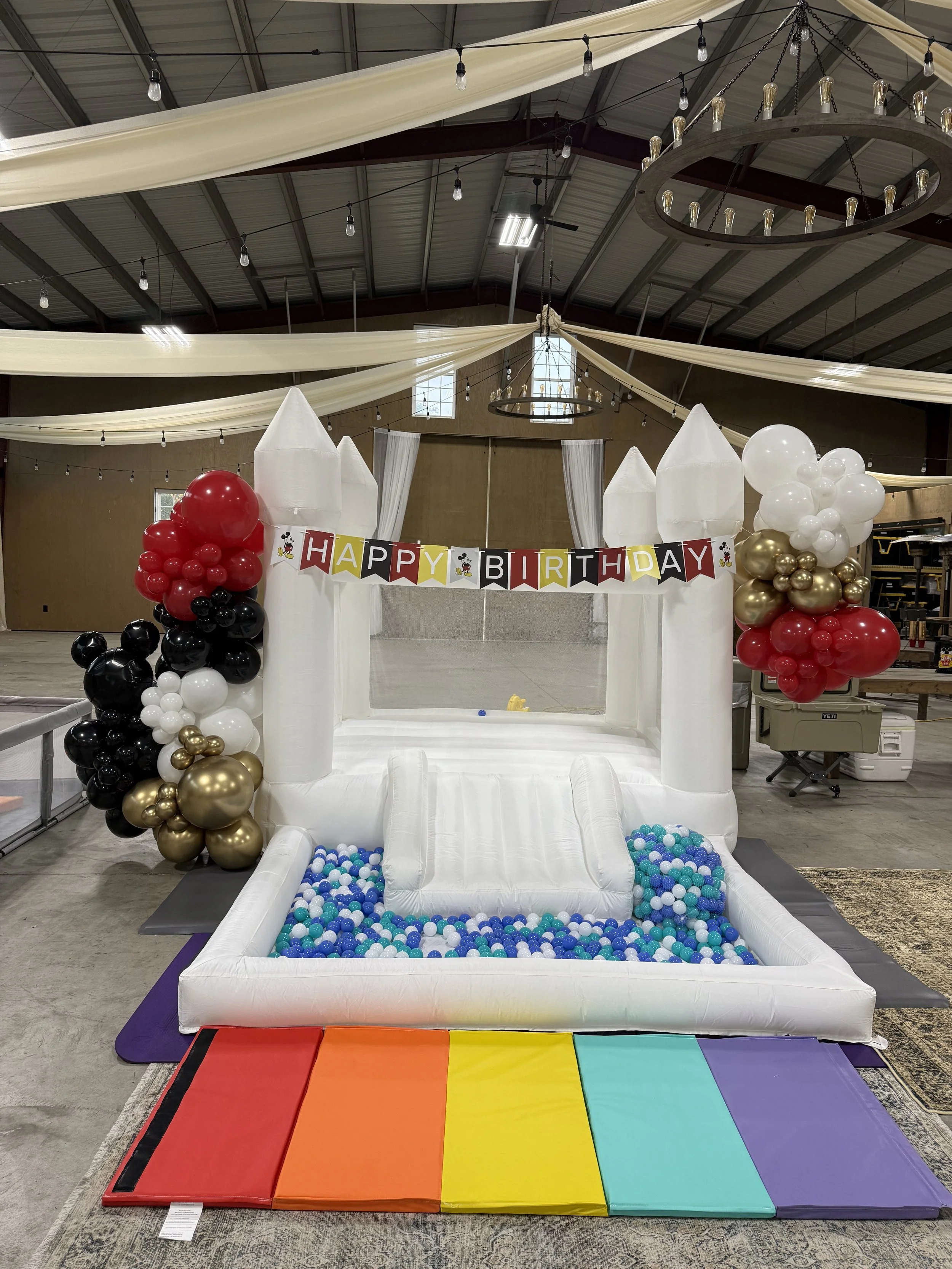 Inflatable white castle with colorful balloons decorating the sides, a rainbow-colored foam mat at the entrance, and a 'Happy Birthday' banner hanging across the front.