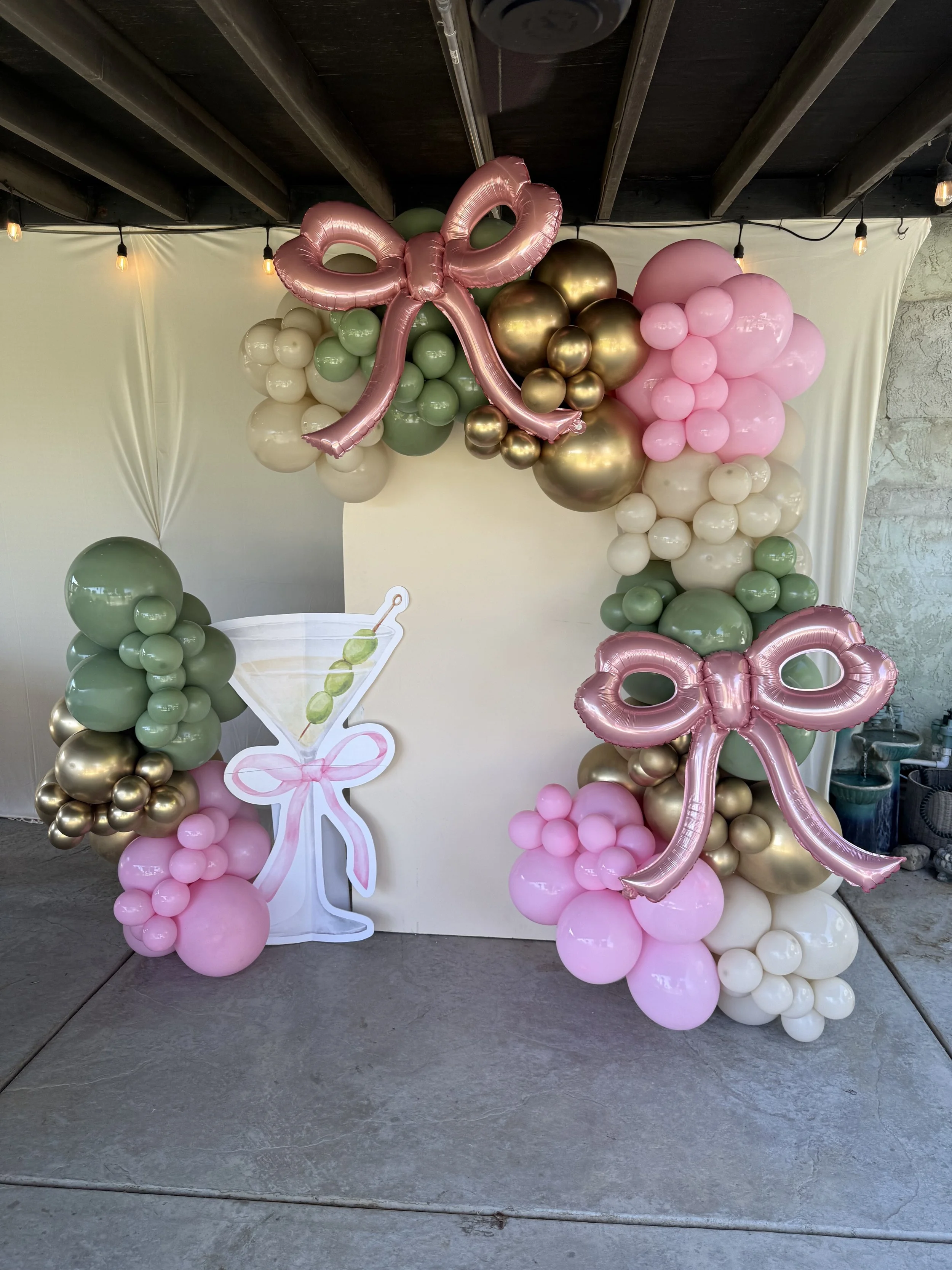 Balloon decoration with pink bows, gold, white, green, and pink balloons arranged in an arch around a white backdrop, with a paper cutout of a martini glass with an olive and pink ribbon,  for a celebration or party.