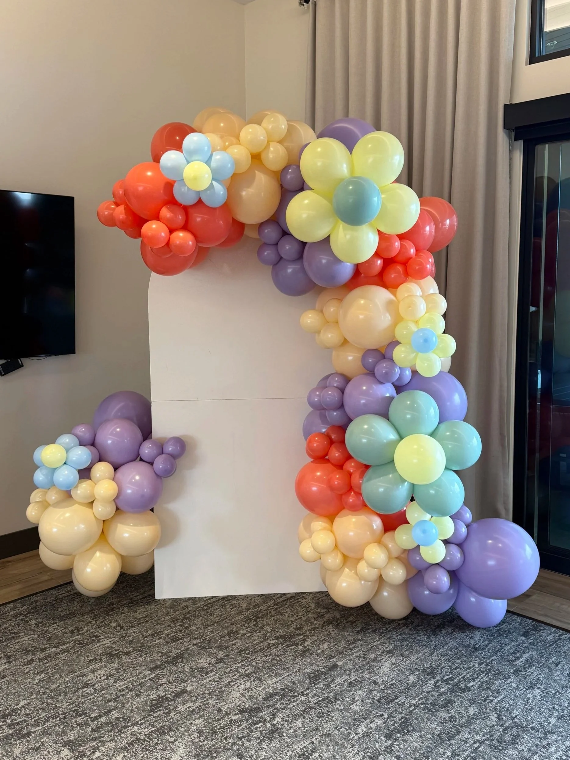 Colorful balloon arrangement with pastel-colored flowers and various sized balloons, forming an arch and cluster against a white backdrop, near a curtain and window.