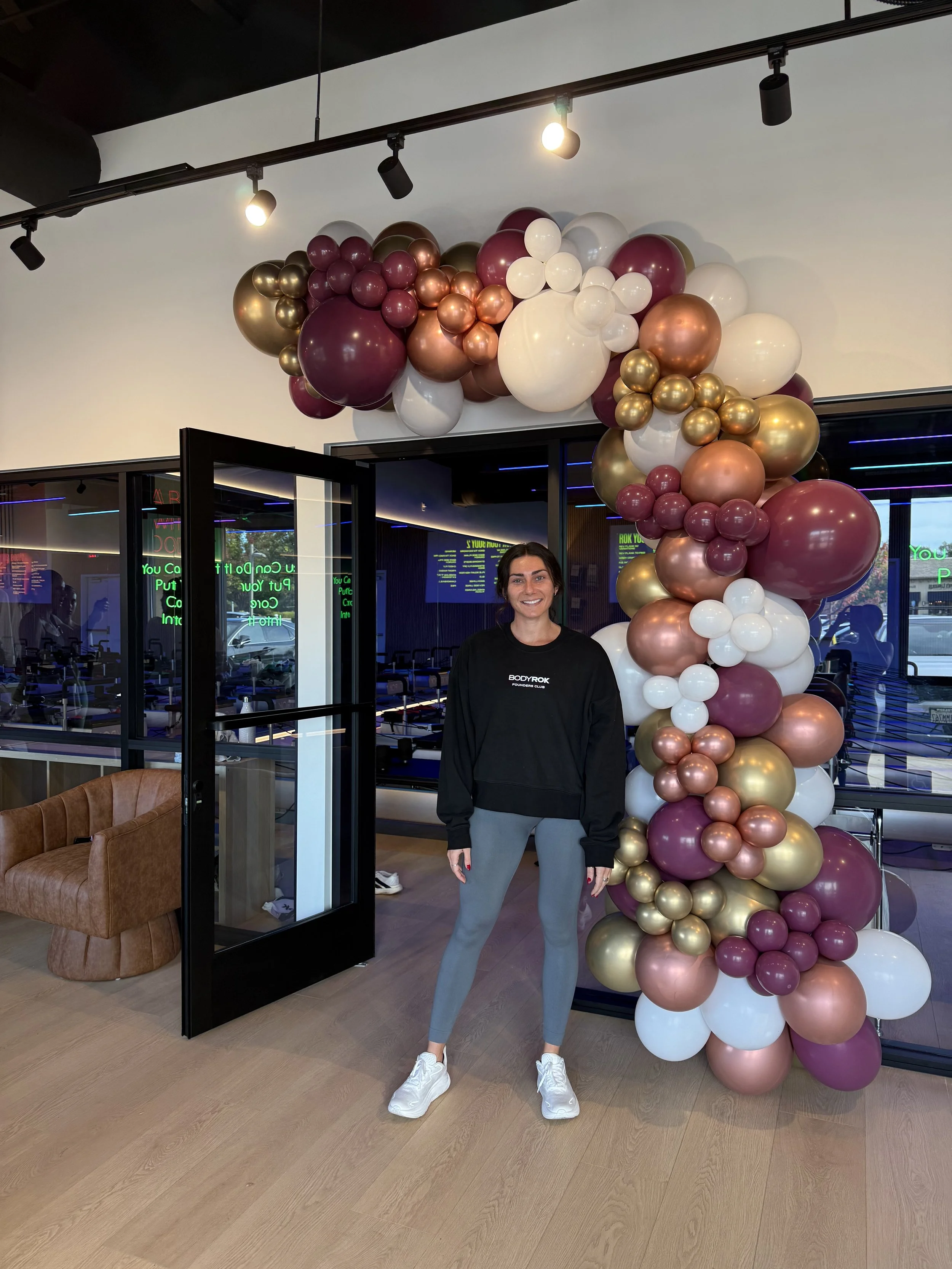 A woman standing in front of a balloon arch at a fitness studio or gym, with a brown armchair and neon lights in the background.