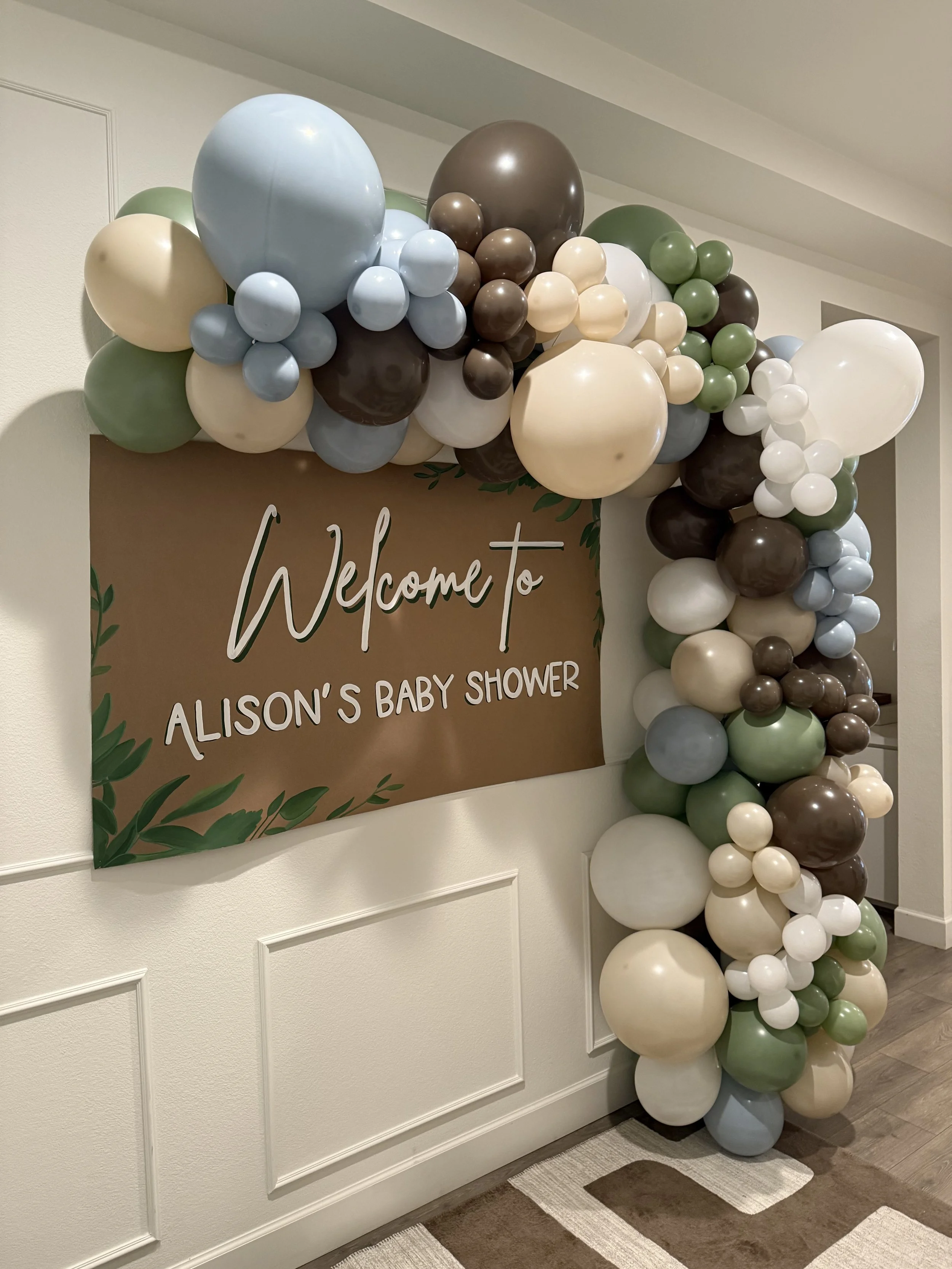 Balloon arch in pastel colors, including light blue, green, cream, white, and brown, framing a brown welcome sign for Alison's baby shower.
