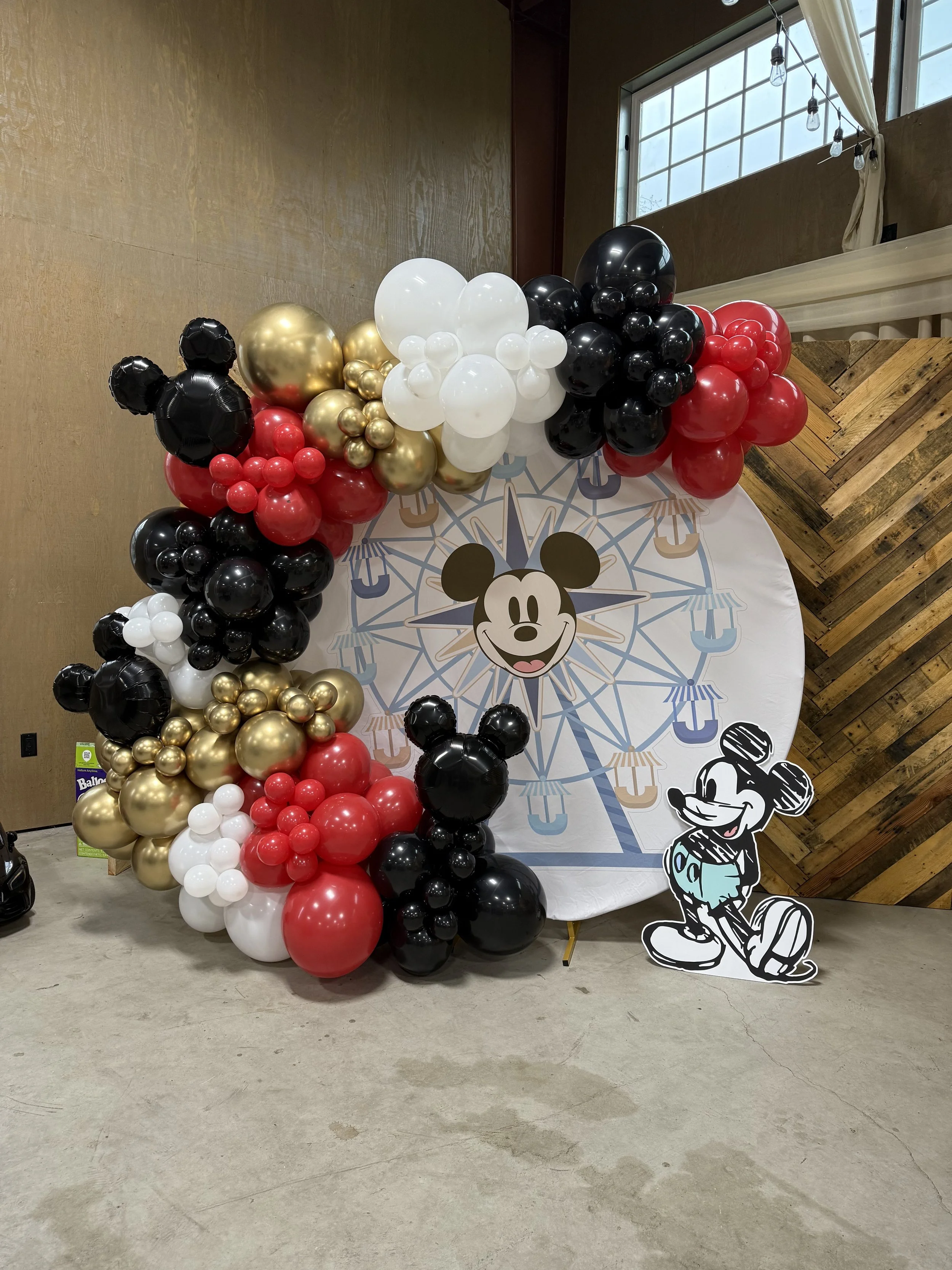 A large balloon arrangement featuring black, white, red, and gold balloons, with Mickey Mouse and Minnie Mouse themed cutouts, set against a backdrop with a Ferris wheel and tent illustrations.