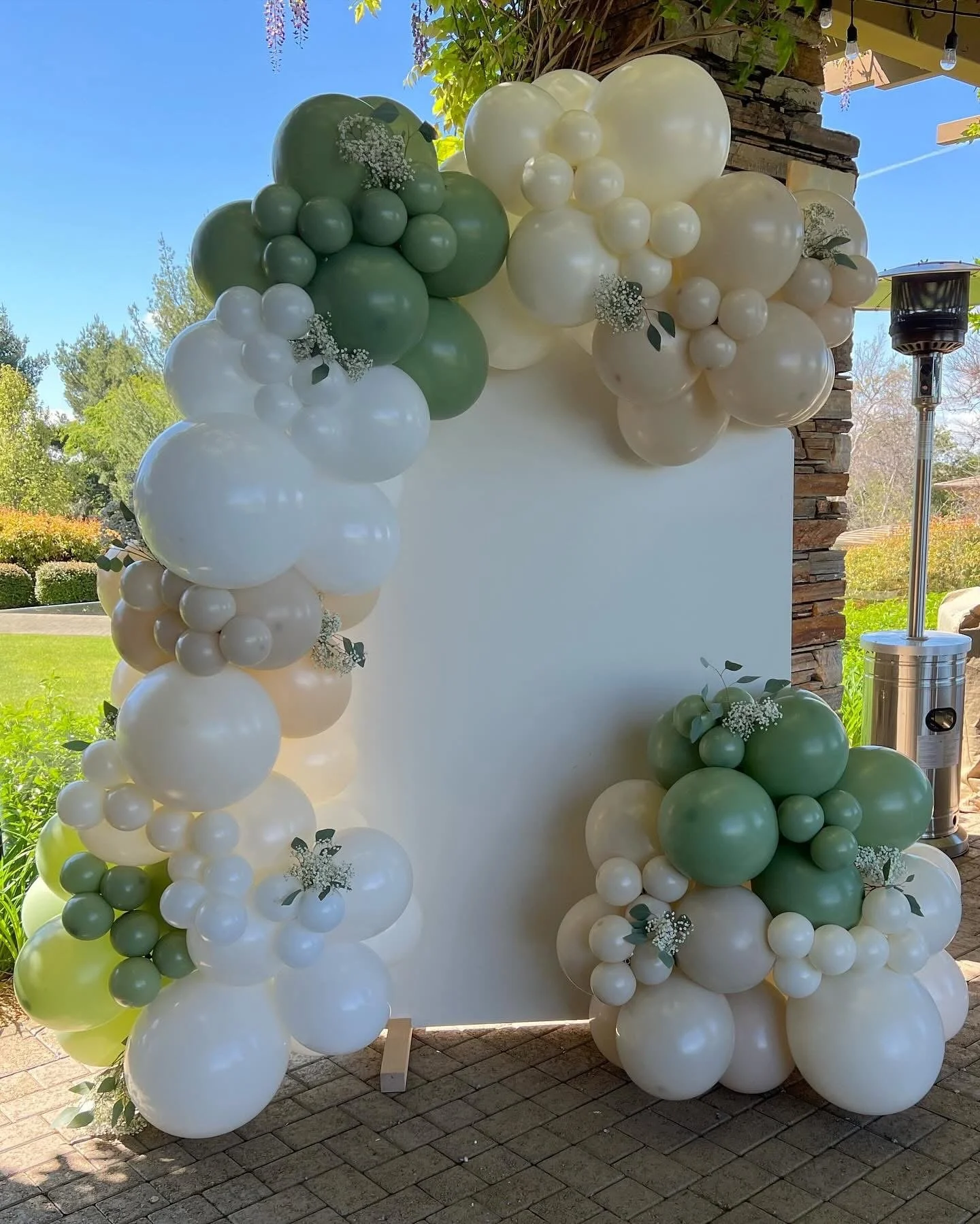 Decorative balloon arch with white, cream, and green balloons, adorned with small white flowers and green leaves, set up outdoors on a sunny day.