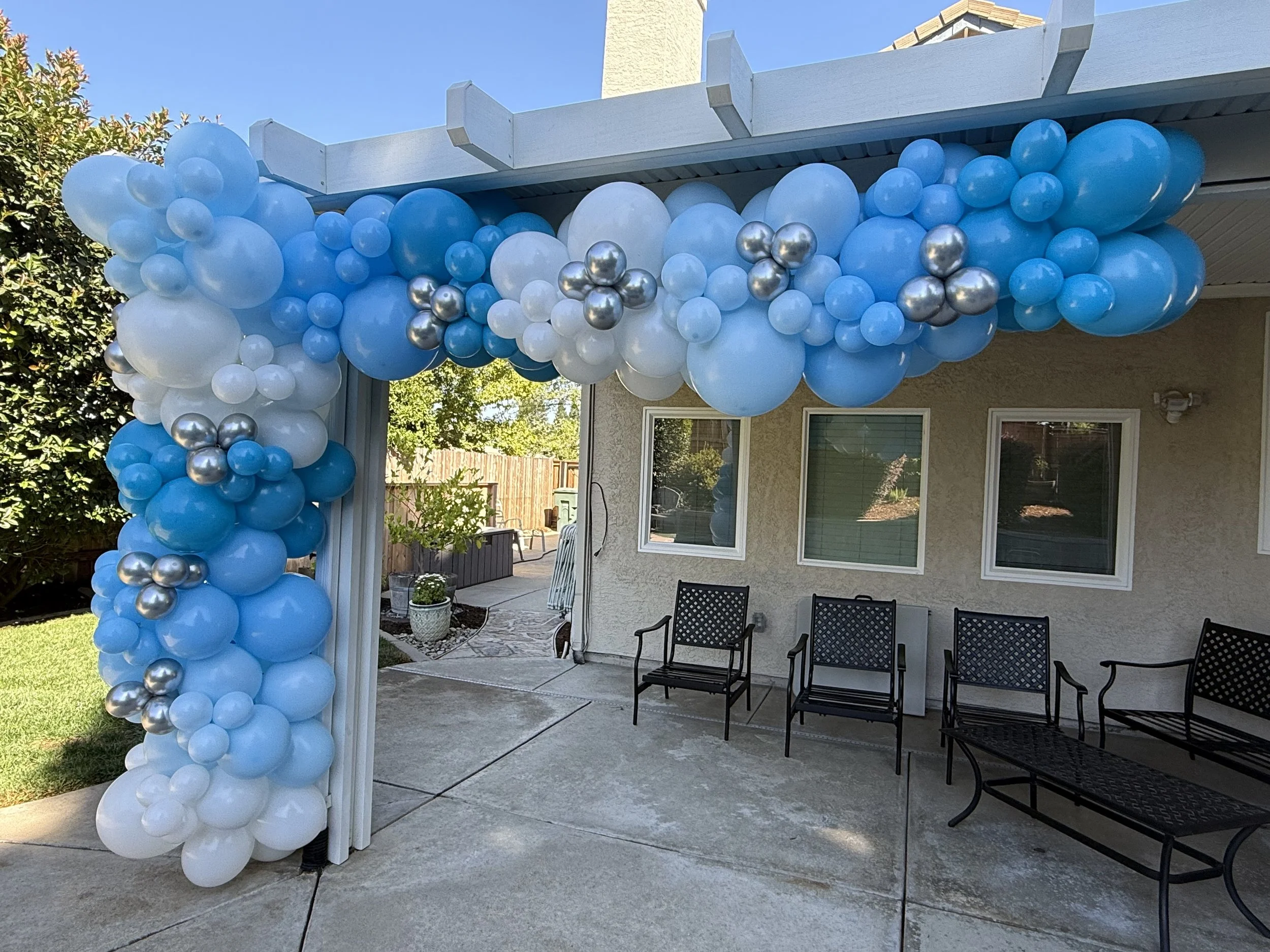 Blue, white, and silver balloons arranged as a decorative arch and column outside a house patio.