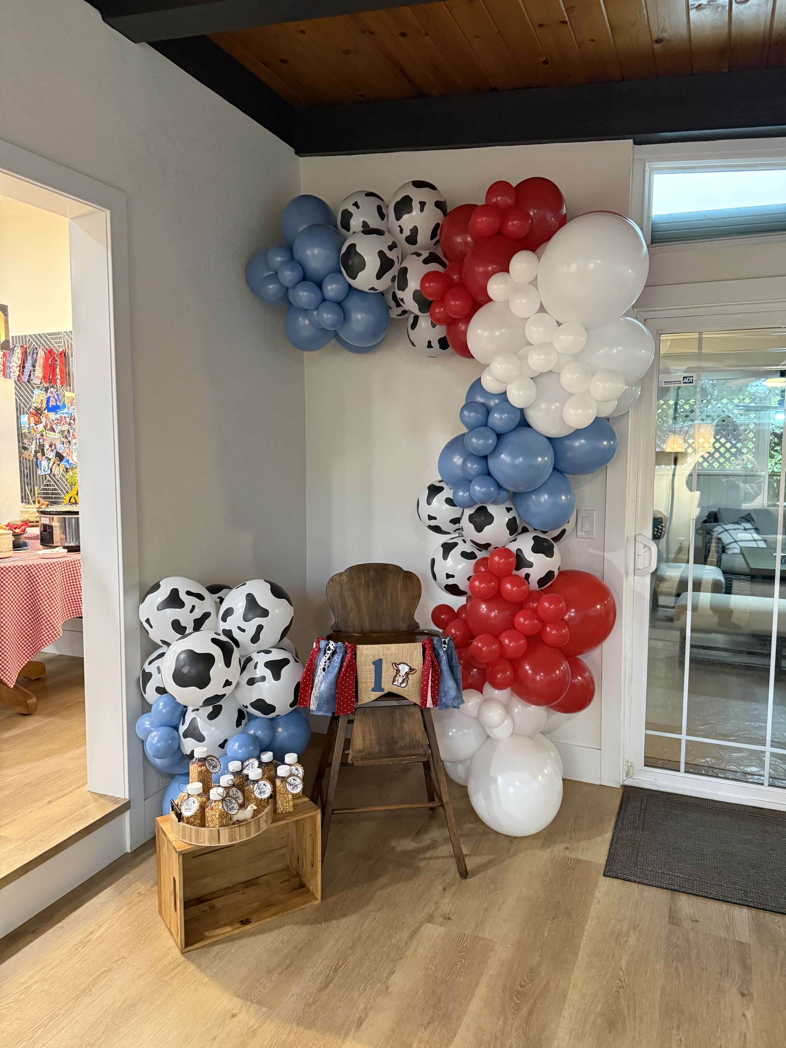 Party decorations with balloon arrangements resembling cow patterns in black and white, red, blue, and white colors, decorated with a sign on a chair and small jars on a wooden stand next to the balloons.