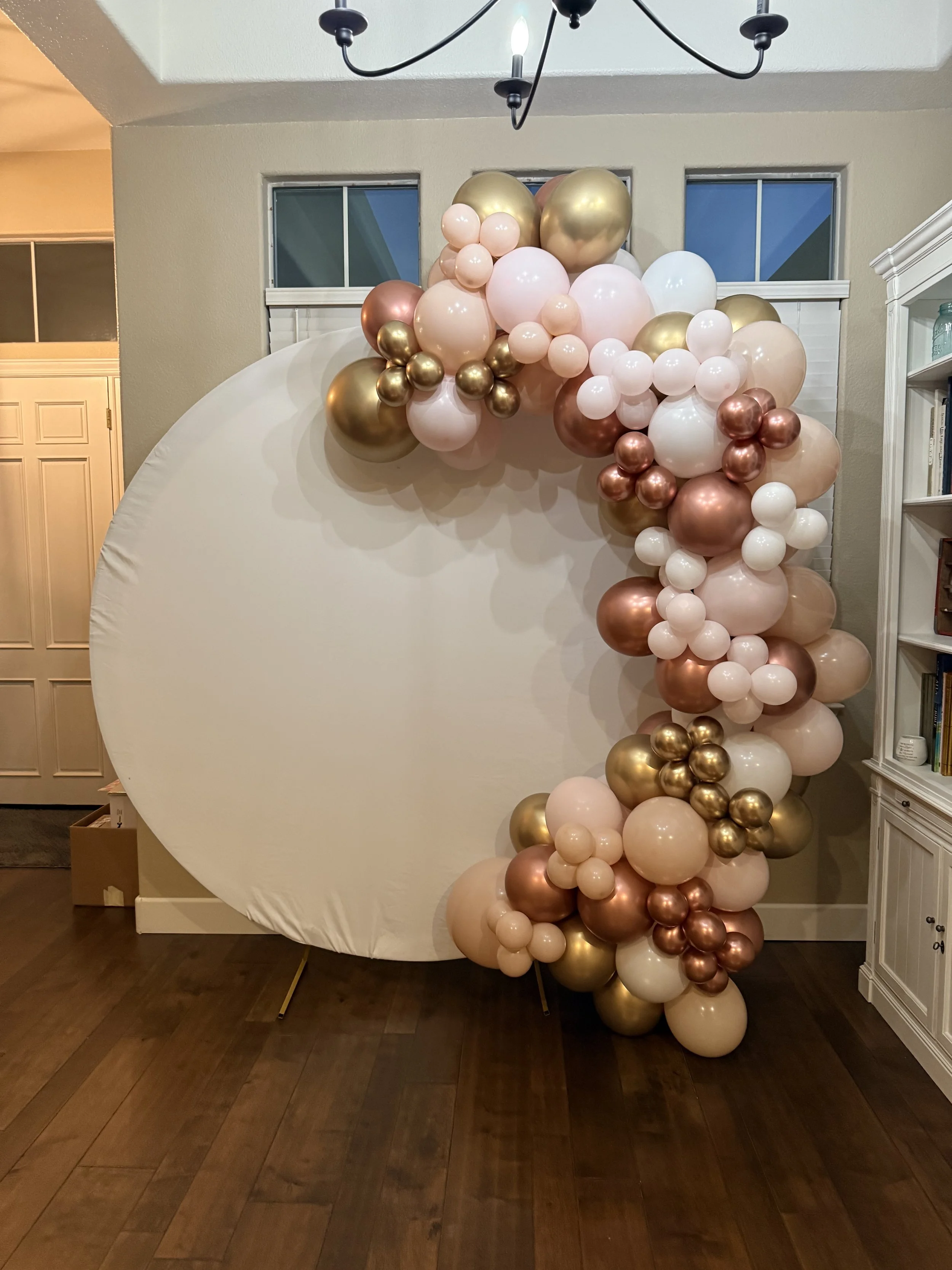 A balloon arch with pink, white, gold, and rose gold balloons arranged around a large white circular backdrop in a decorated indoor setting.