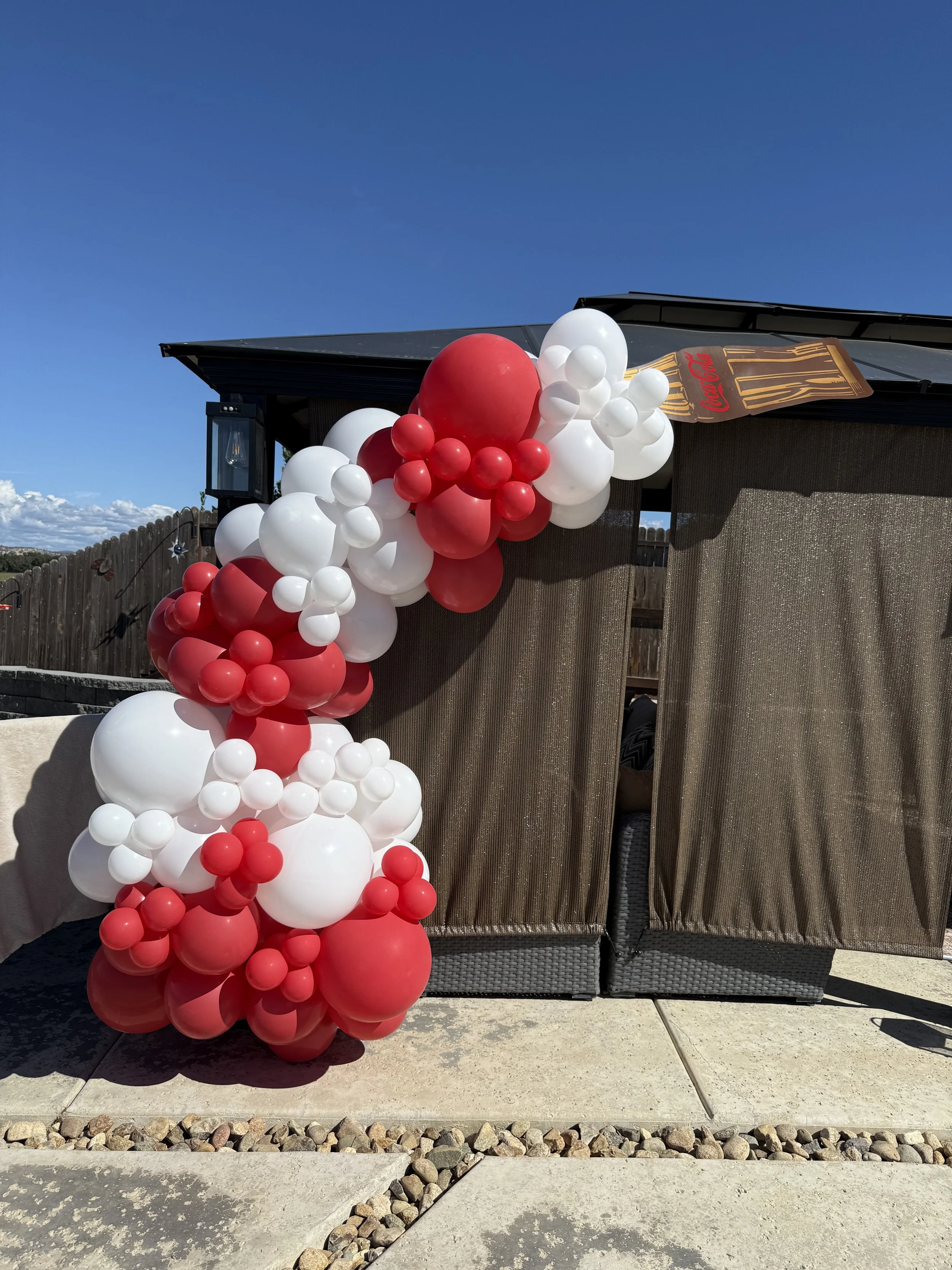 Decorative balloon arch with red, white, and pink balloons outside a wicker gazebo with brown curtains on a sunny day.