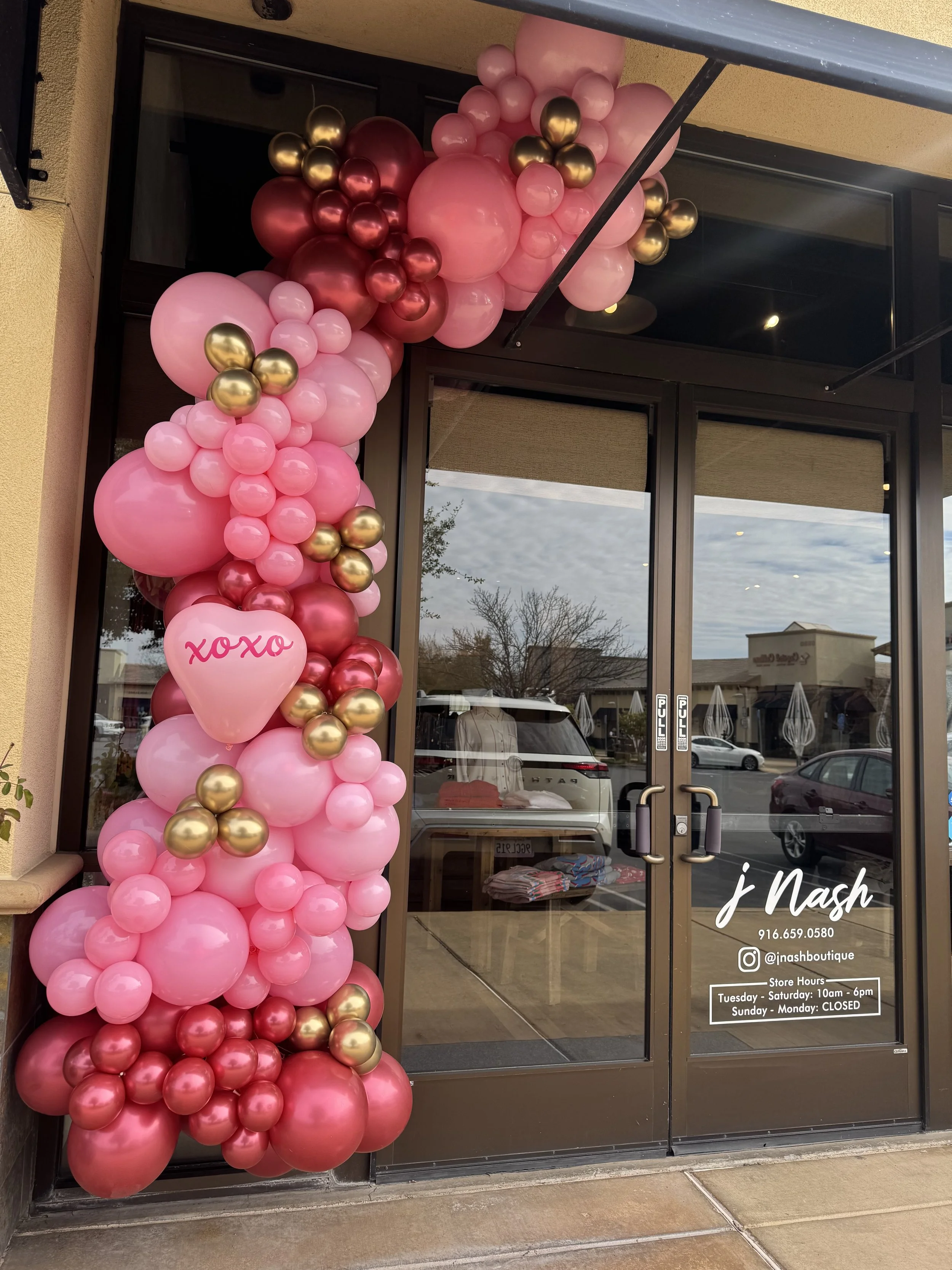 Decorative pink, red, and gold balloons arranged along the side of a store entrance door with a pink heart-shaped balloon that has 'XOXO' written on it. The store is called 'j Nash' and provides contact information, social media, and business hours o