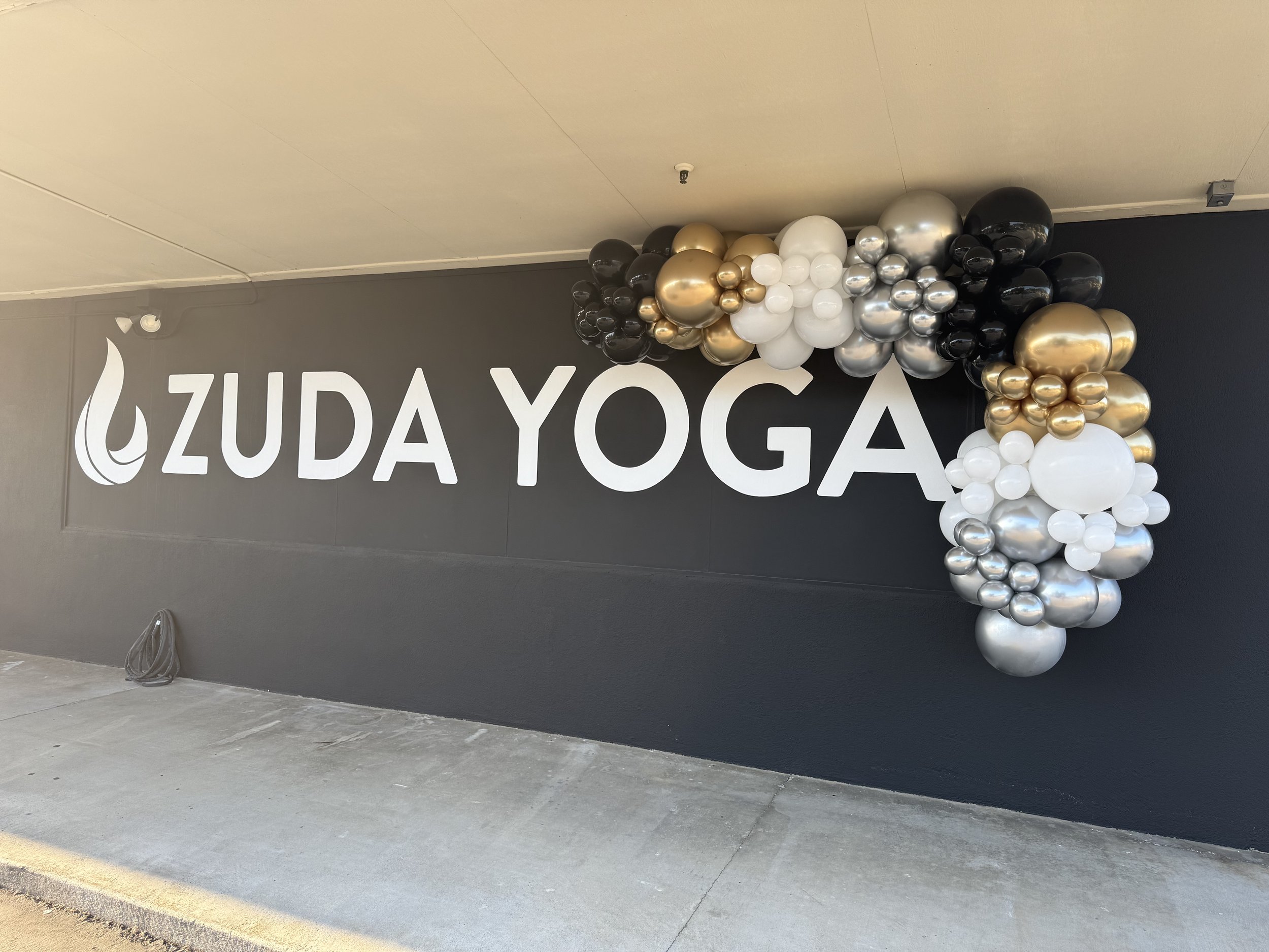 A black wall with large white letters spelling 'ZUDA YOGA' and a decorative arrangement of black, white, gold, and silver balloons in a cluster on the right side.