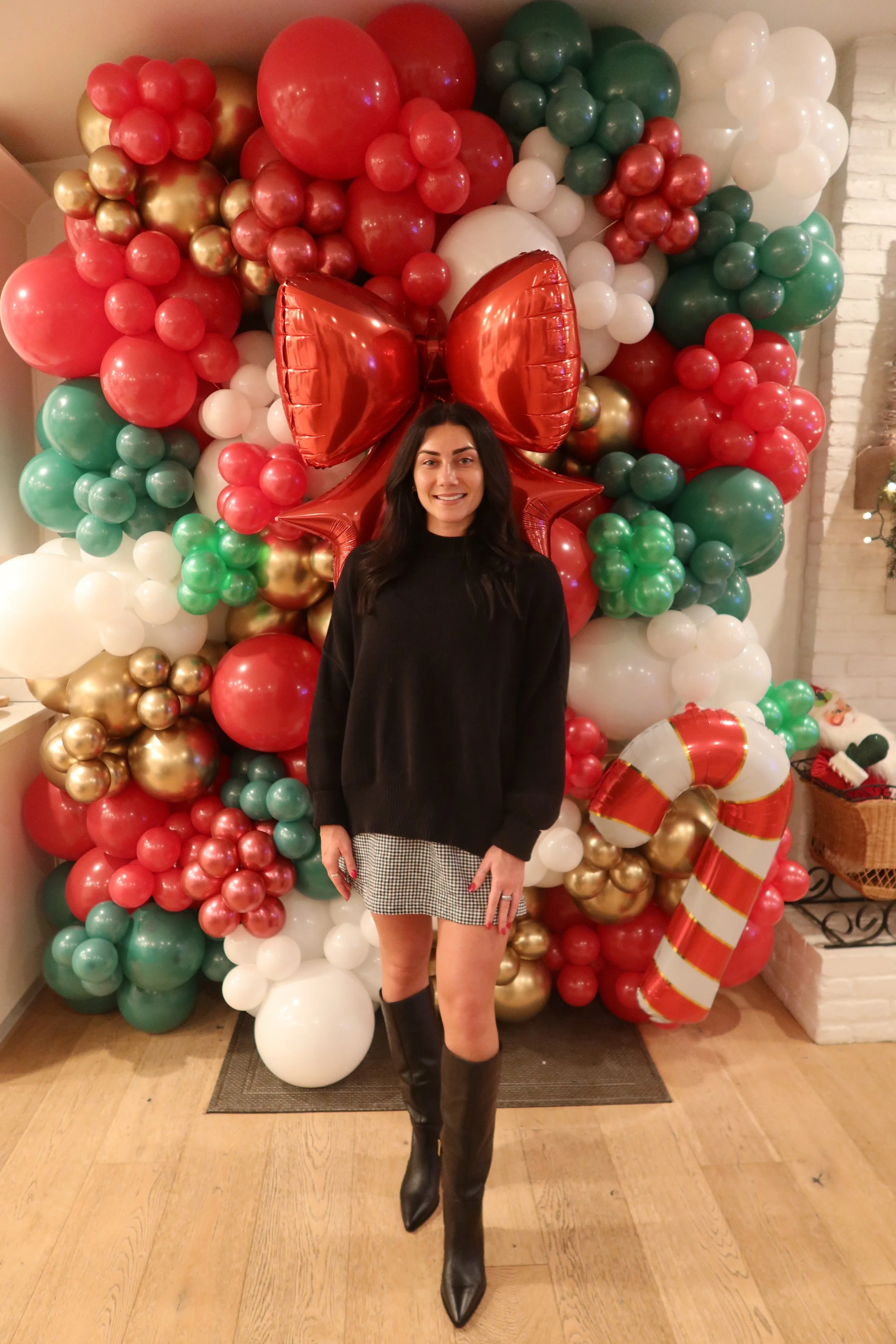A woman standing in front of a Christmas balloon decoration, wearing a black sweater, checkered skirt, and black knee-high boots.