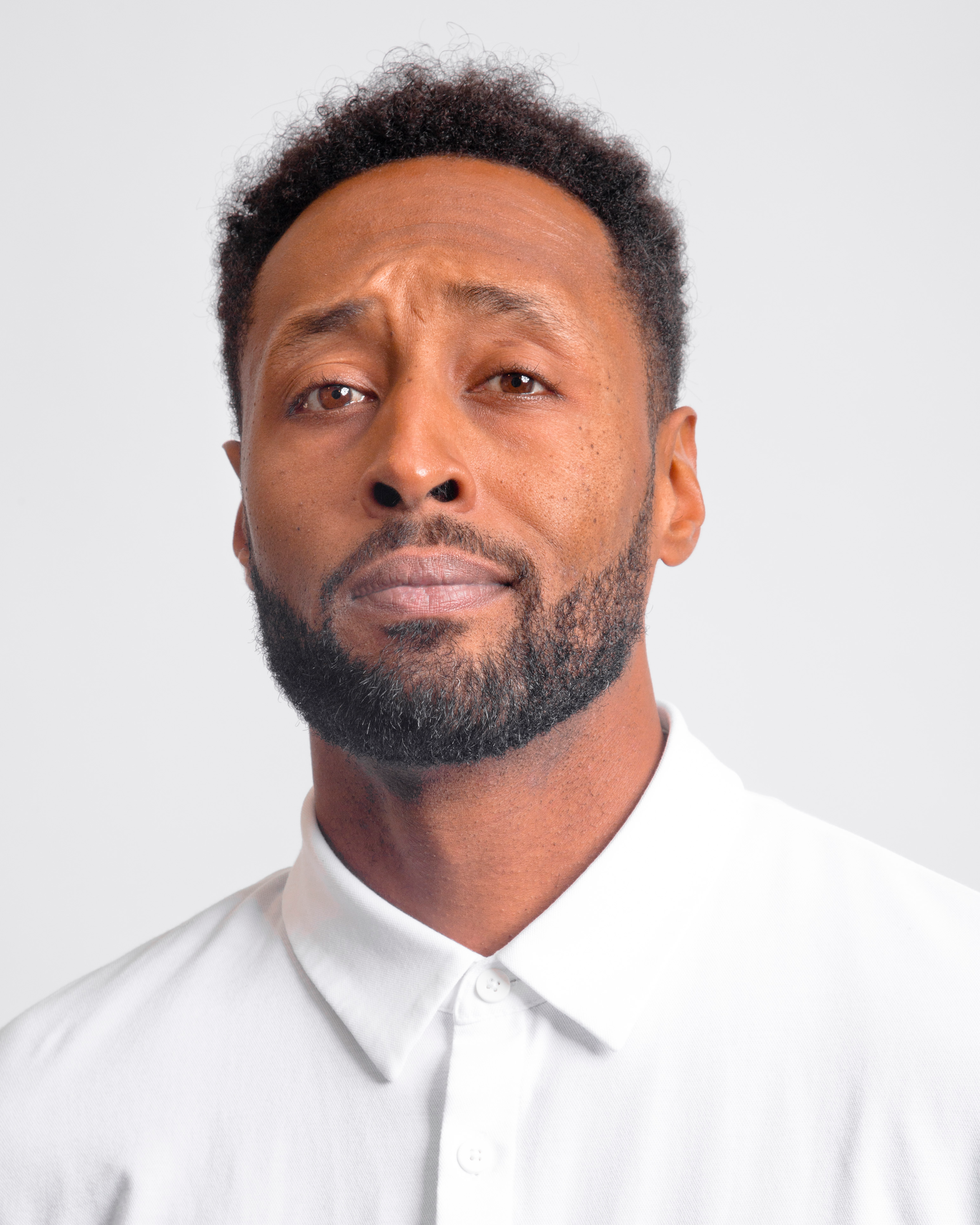 Close-up portrait of a man with a beard and short curly hair, wearing a white collared shirt, against a plain white background.