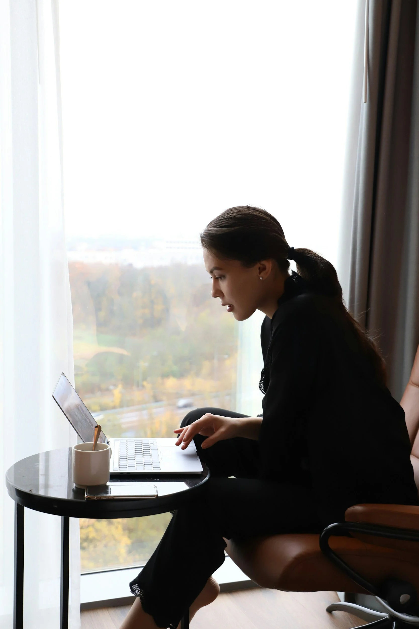 Young woman with dark hair in a ponytail sitting on an office chair by a small round table, looking at a laptop screen, with a white mug and a smartphone on the table, in front of a large window with sheer curtains and a city view in the background.