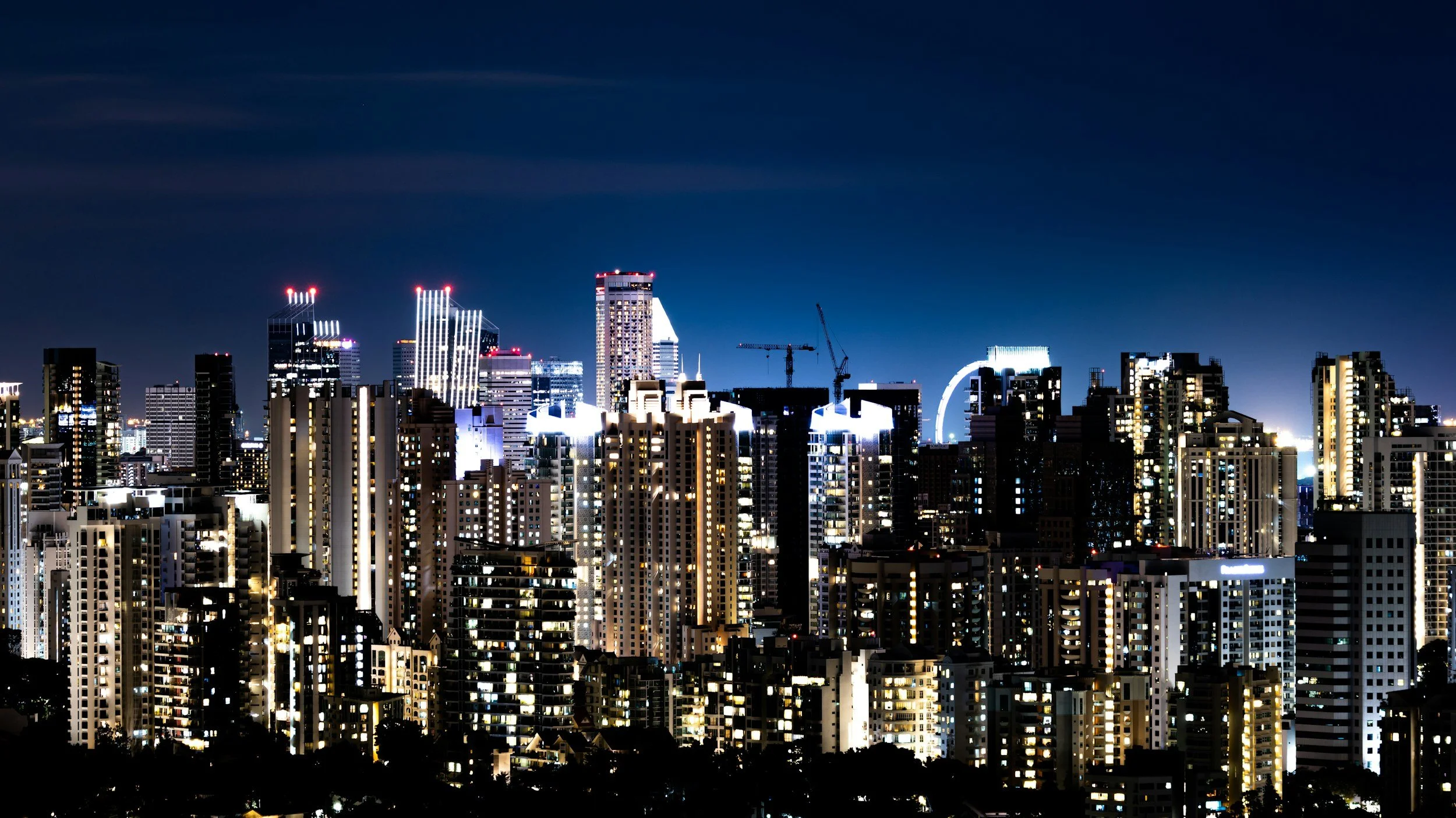 Nighttime city skyline with illuminated high-rise buildings and construction cranes against a dark sky.