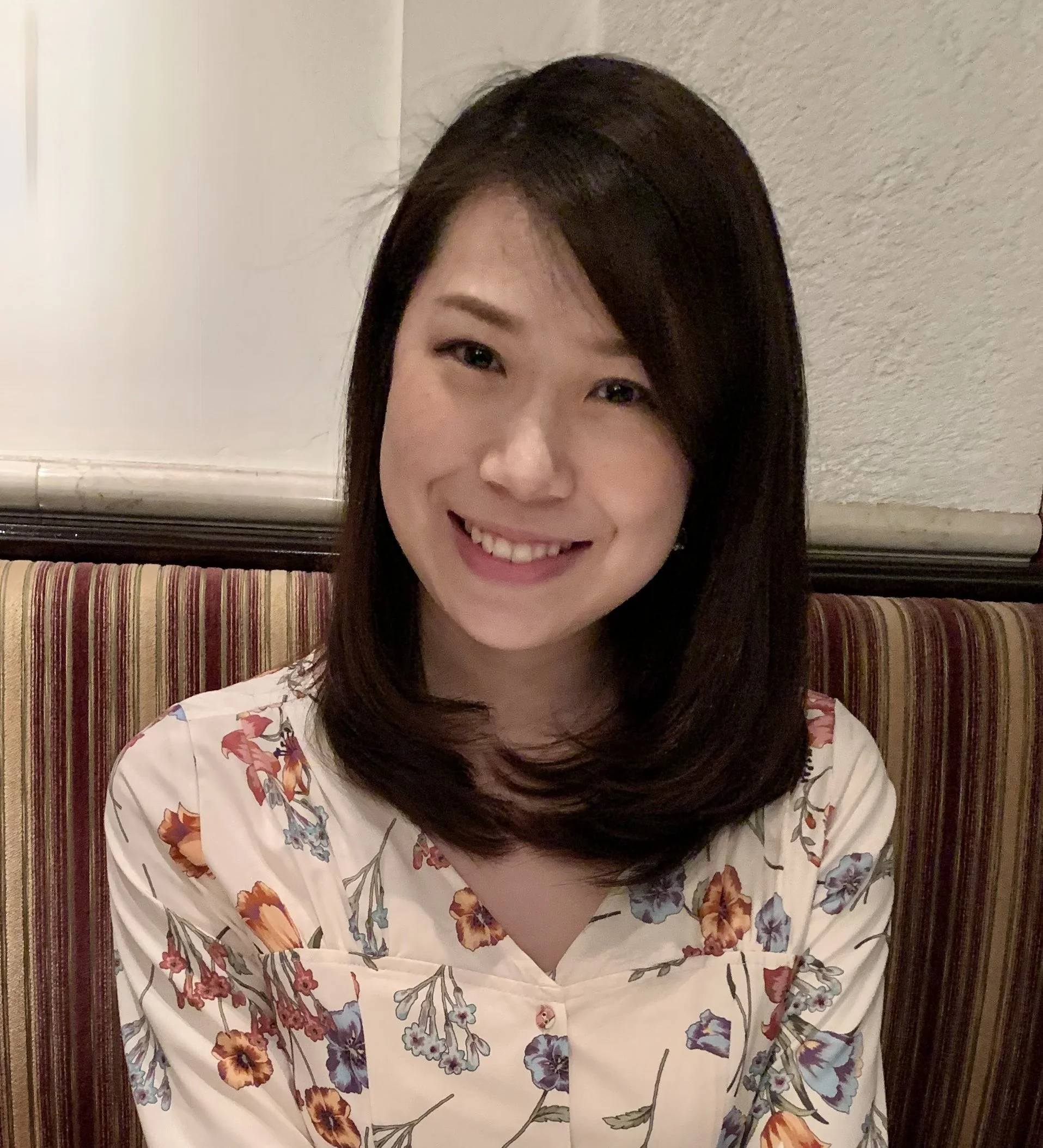 A young Asian woman with shoulder-length dark hair smiling, wearing a floral-patterned blouse, sitting on a striped booth in a restaurant or cafe.