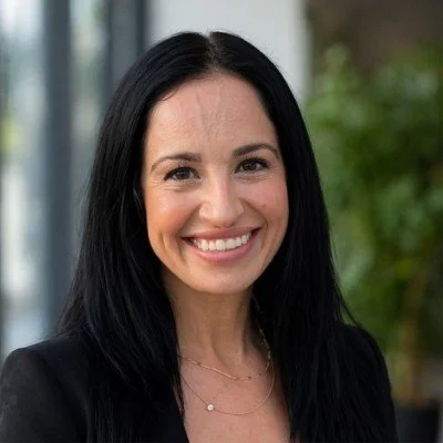 A woman smiling with long black hair, wearing a black blazer and layered necklaces, standing outdoors with greenery in the background.