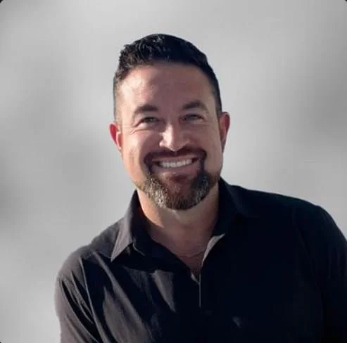 Smiling man with short dark hair and beard, wearing a black shirt, standing against a plain light background.