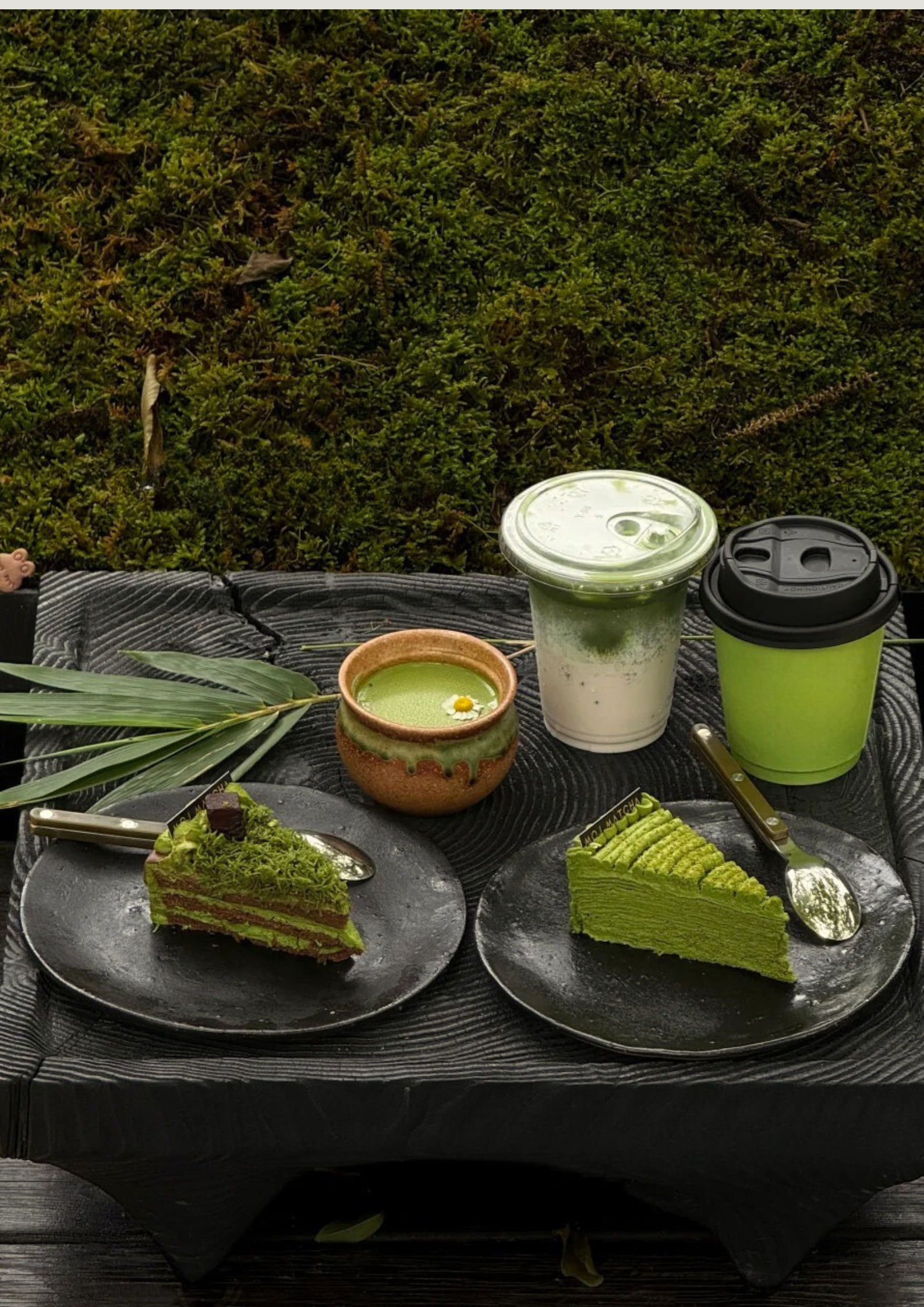 Two slices of matcha green tea cake on black plates, a green matcha latte, a green matcha drink in a to-go cup, and a green matcha drink in a plastic cup with a lid, all placed on a black tray outside on a wooden surface with greenery in the background.