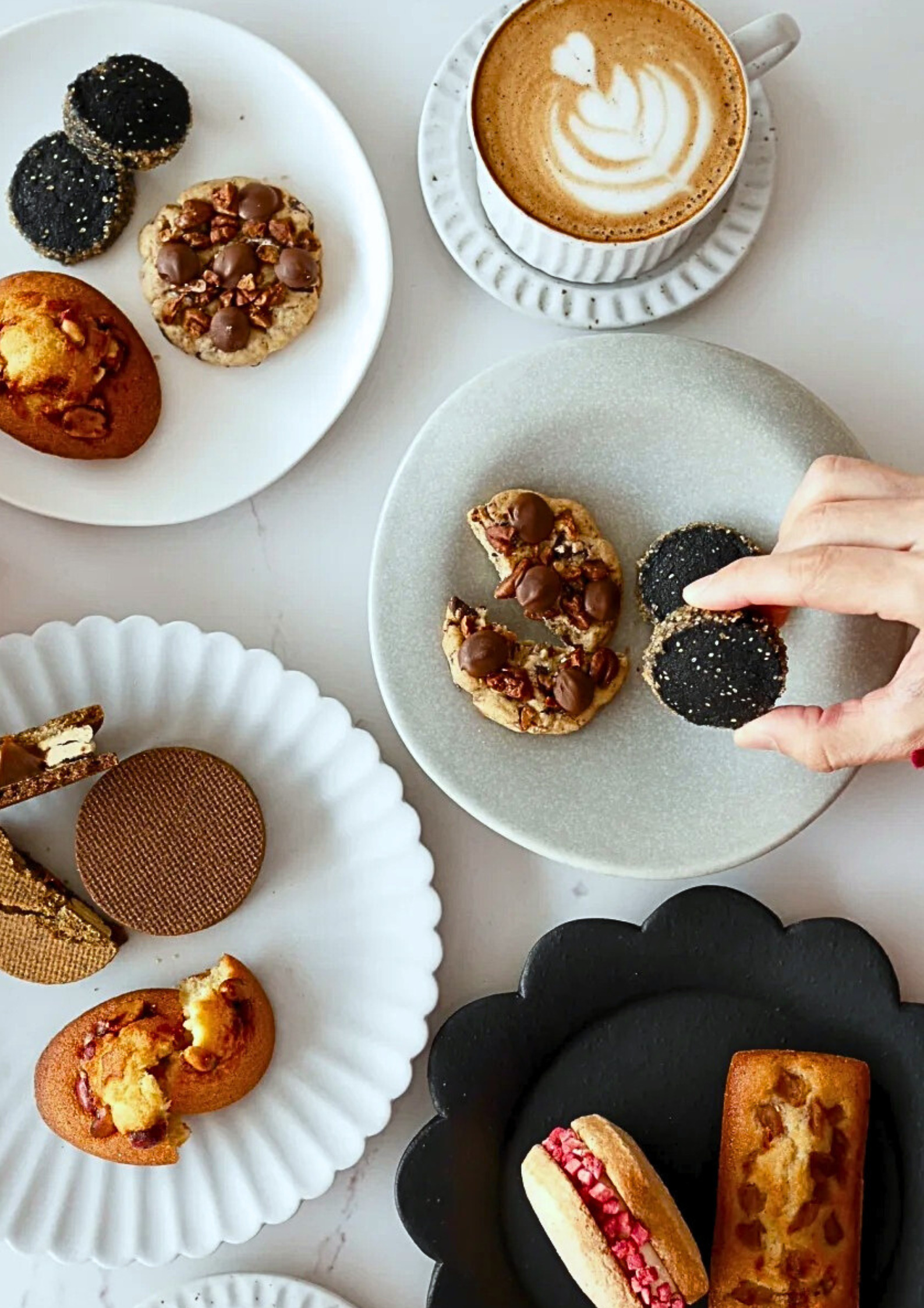 A table with an assortment of baked goods and a cup of latte with foam art. There are cookies with nuts and chocolate, macarons, and other pastries on various plates.