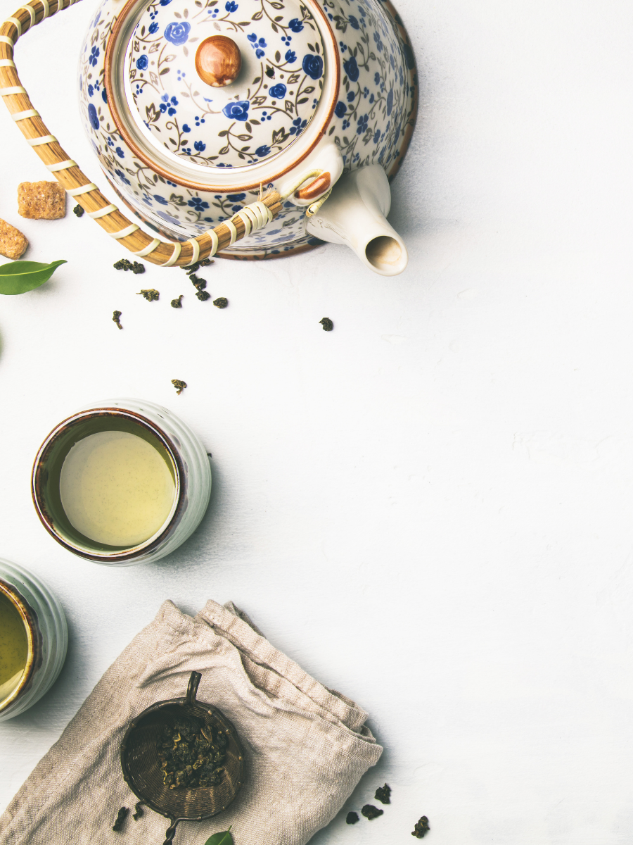 A floral patterned teapot with a bamboo handle, two cups of green tea, loose tea leaves in a tea infuser on a beige cloth, and scattered tea leaves on a white surface.