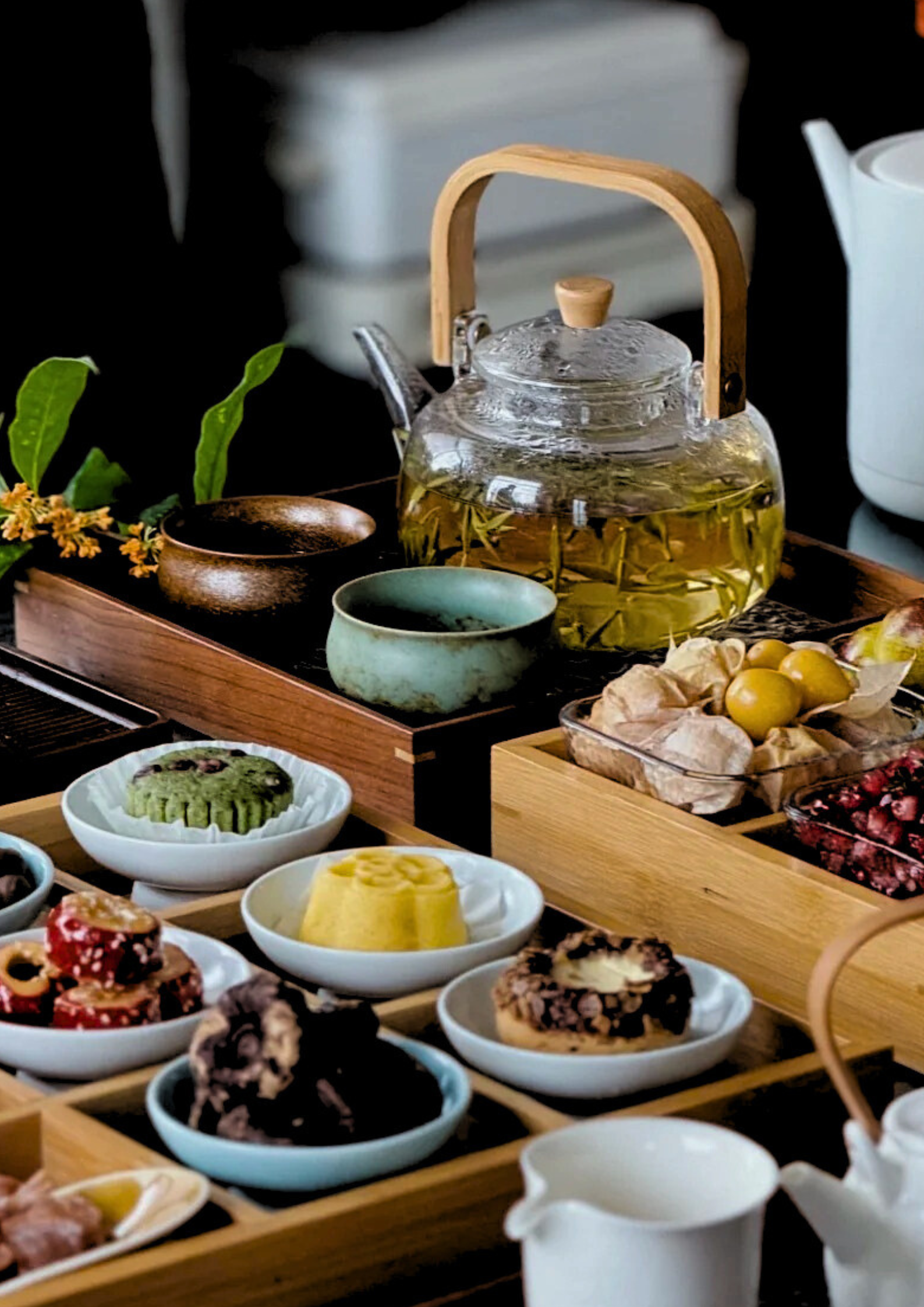 Tea set with a glass teapot, cups, and various desserts and snacks on a wooden tray and bowls, with green leaves and flowers nearby.