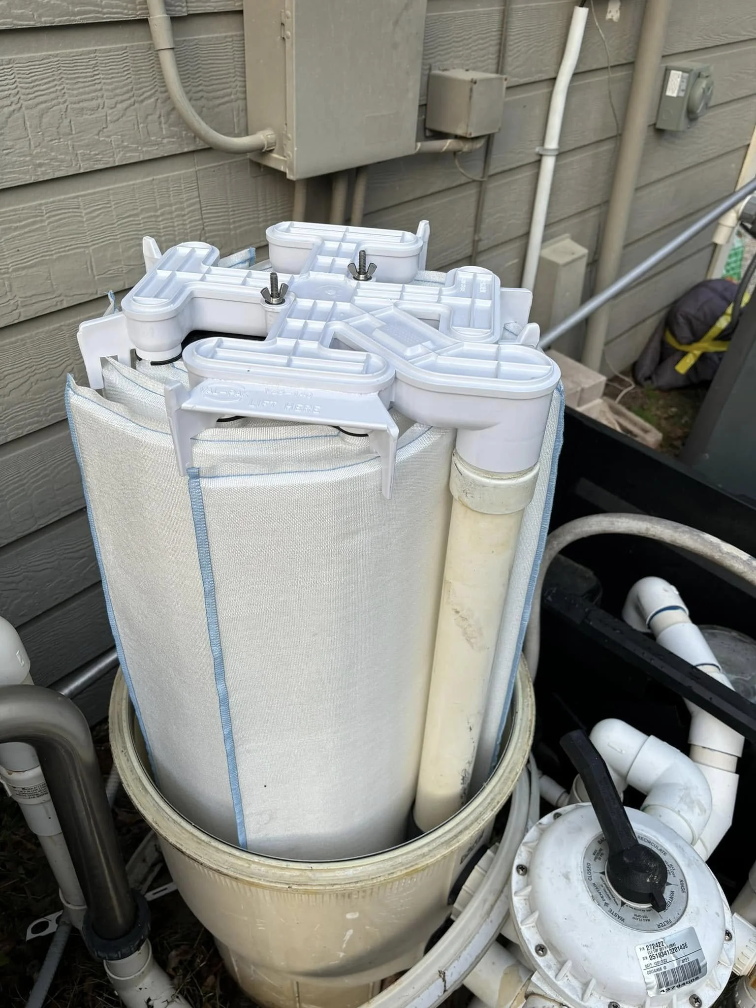 Pool filtration system parts including a large white cartridge filter, pipes, and a pump, installed outside a house with siding.