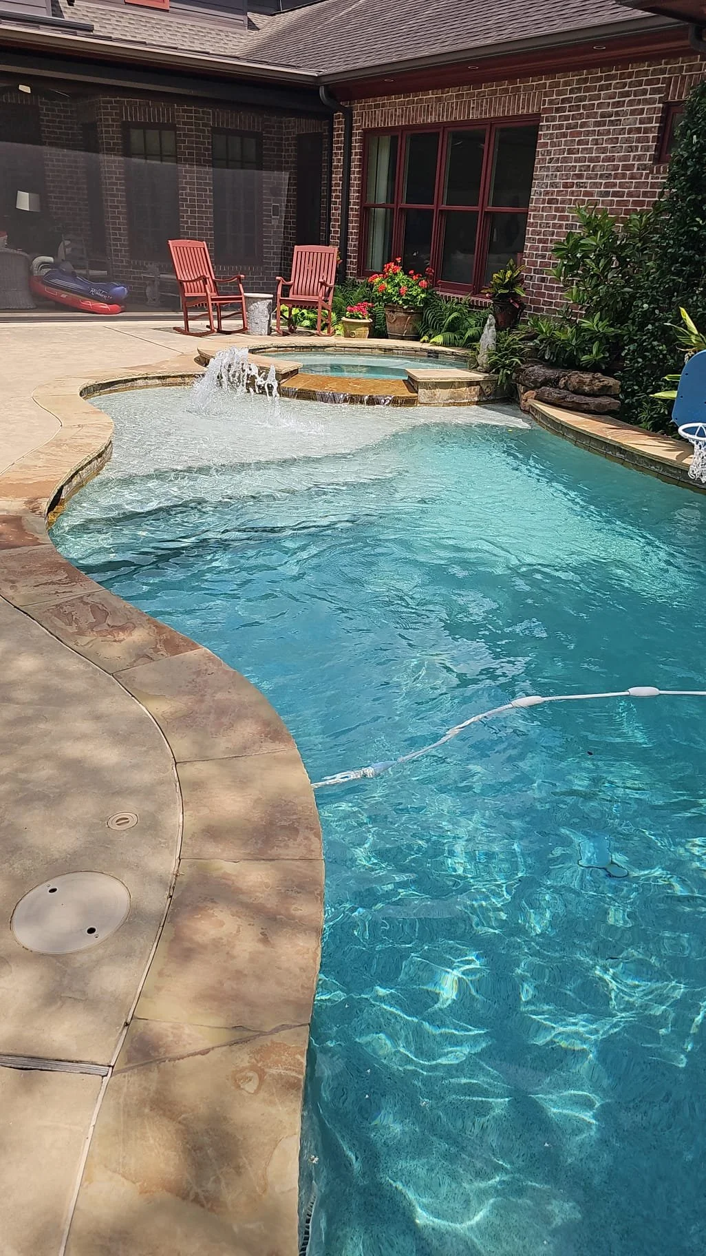 A backyard swimming pool with a hot tub, surrounded by a concrete deck and garden plants. There are two red chairs near the house's brick wall, which has large windows and flower pots. The swimming pool has a fountain and a pool cleaner floating in the water.