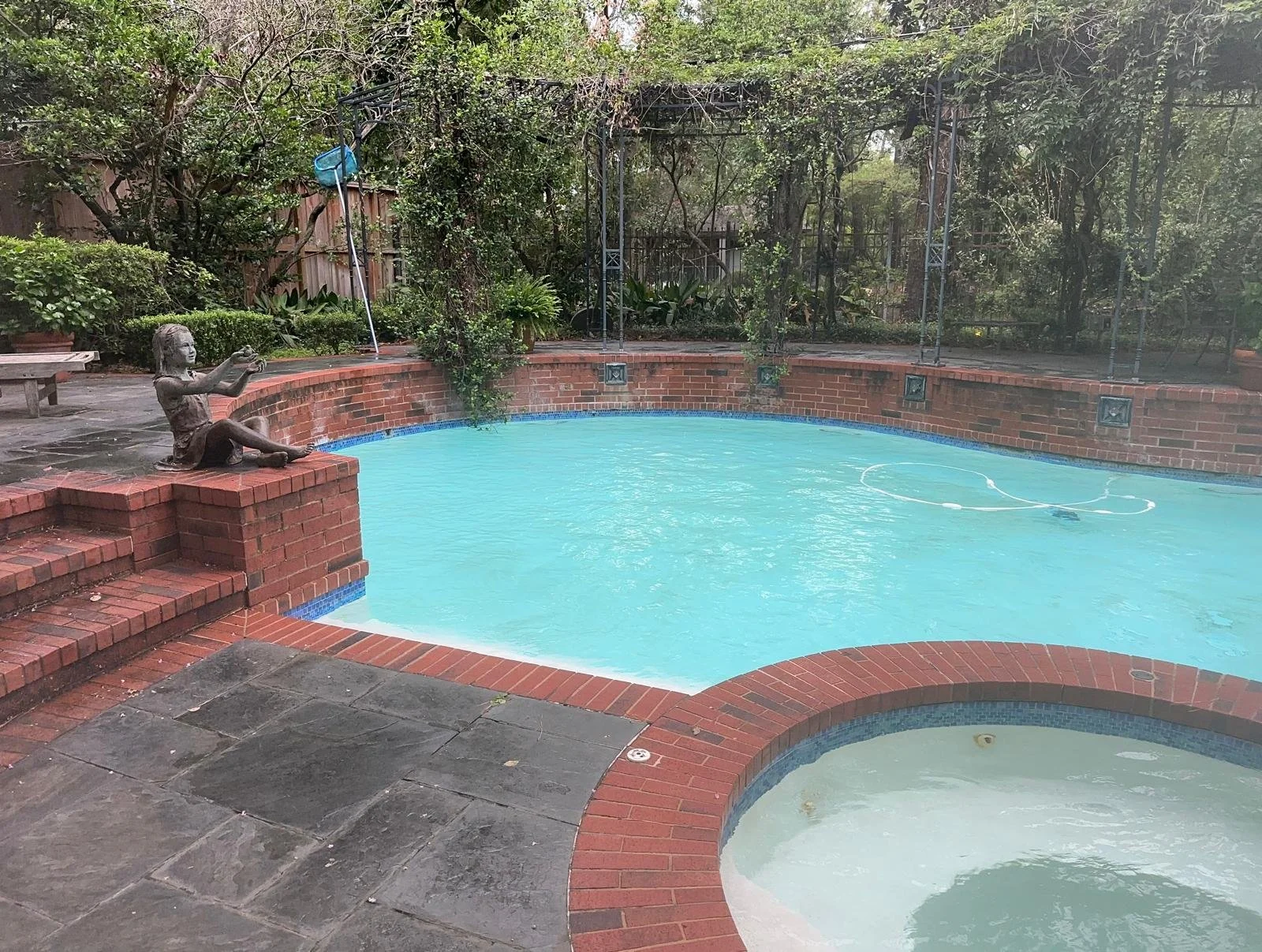 An outdoor swimming pool with a small attached hot tub, surrounded by a brick patio. There is a statue of a girl sitting cross-legged, holding a small object, featuring on the ledge of the pool. The background has trees and greenery, with a metal fence enclosure and pool cleaning equipment visible.