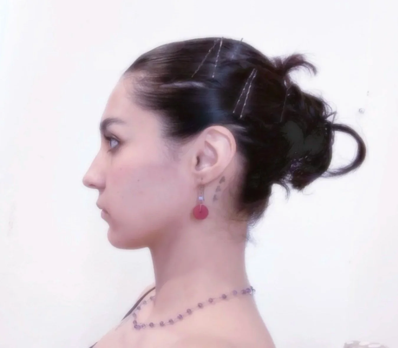 A side profile of a woman with dark brown hair styled in an updo with hairpins, wearing earrings, a beaded necklace, and makeup with pink blush.