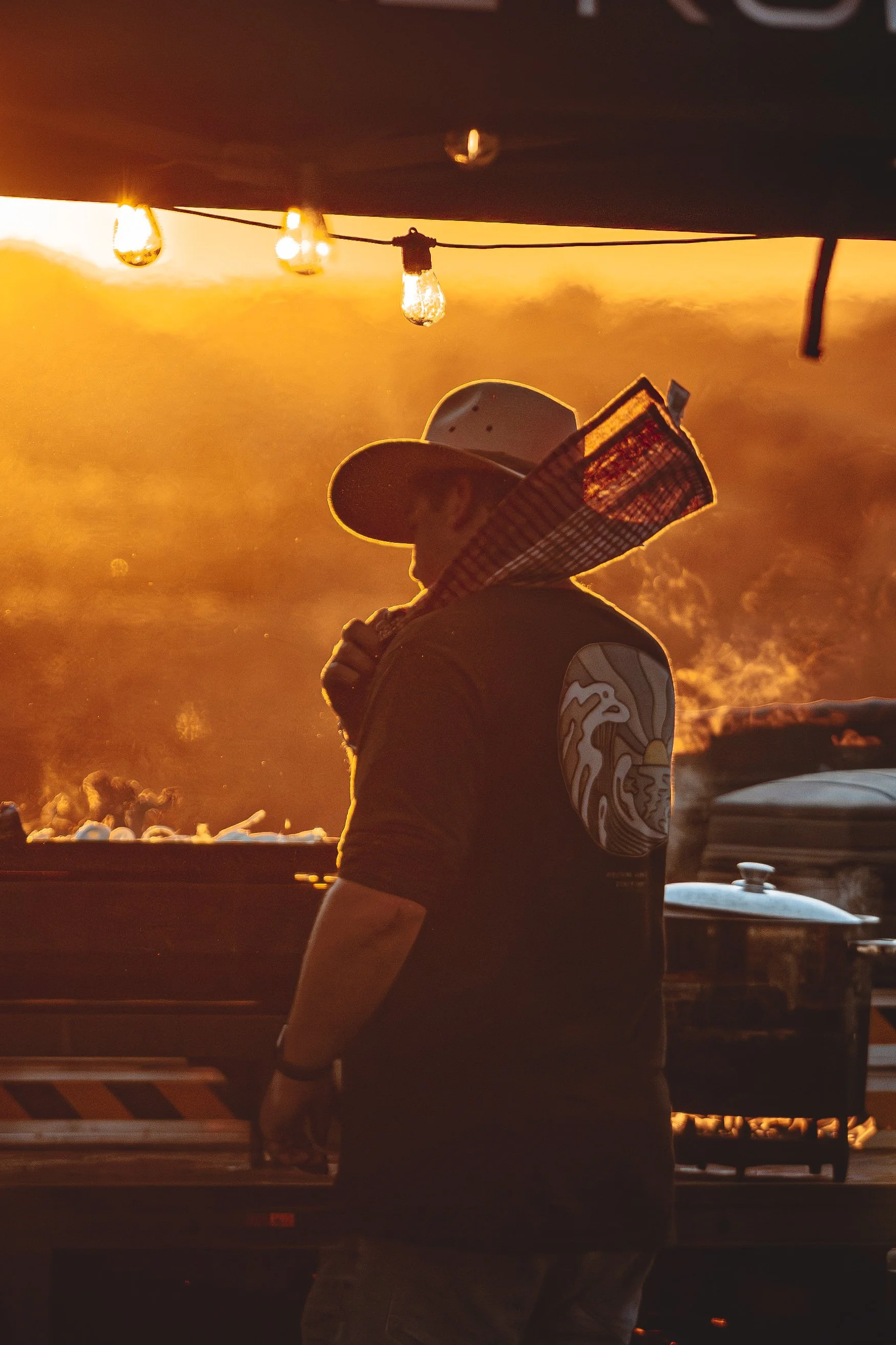 A person wearing a hat and sunglasses standing by a grill with smoke at sunset, decorated with string lights.