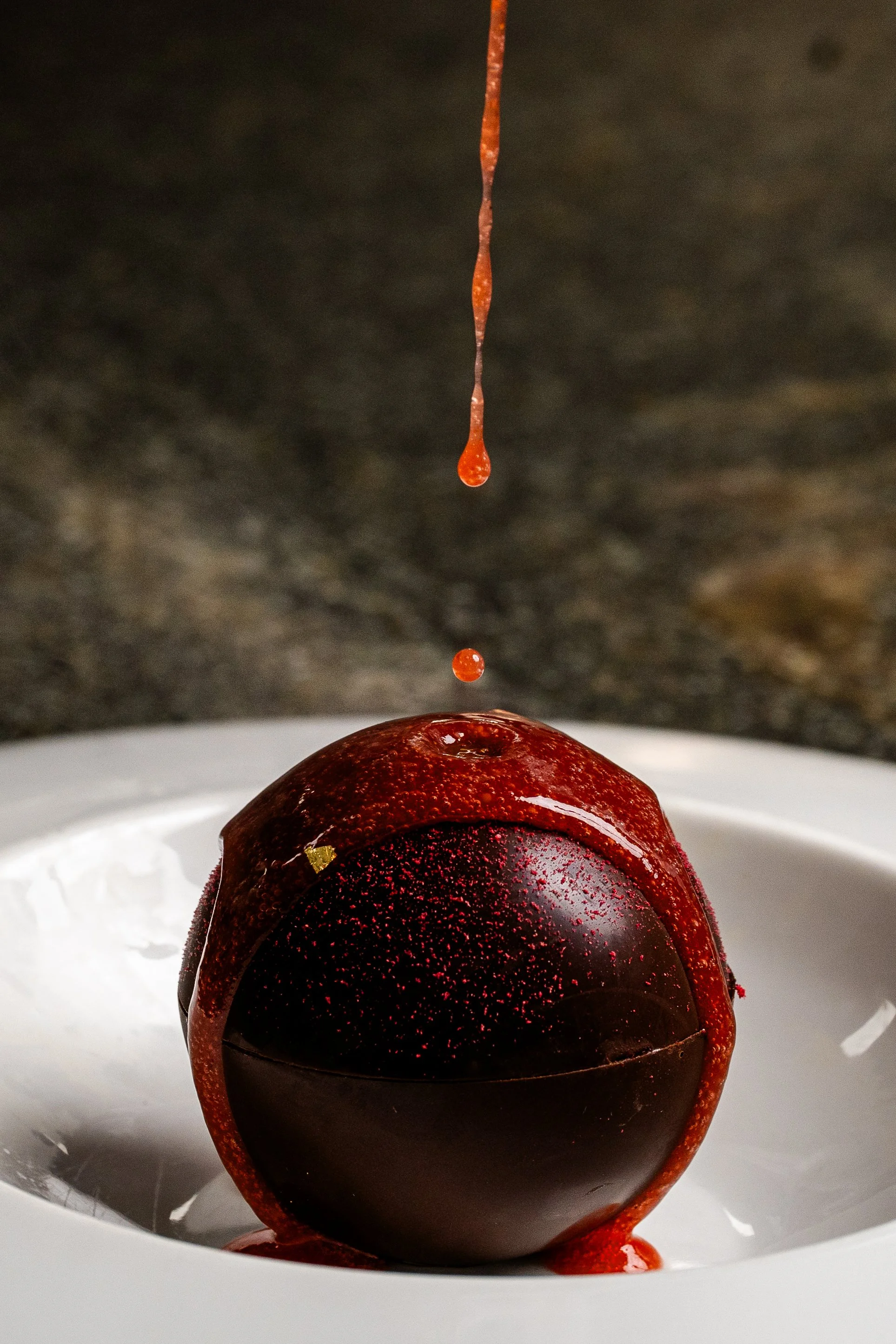 Close-up of a dark chocolate sphere on a white plate, with red sauce dripping onto it from above.