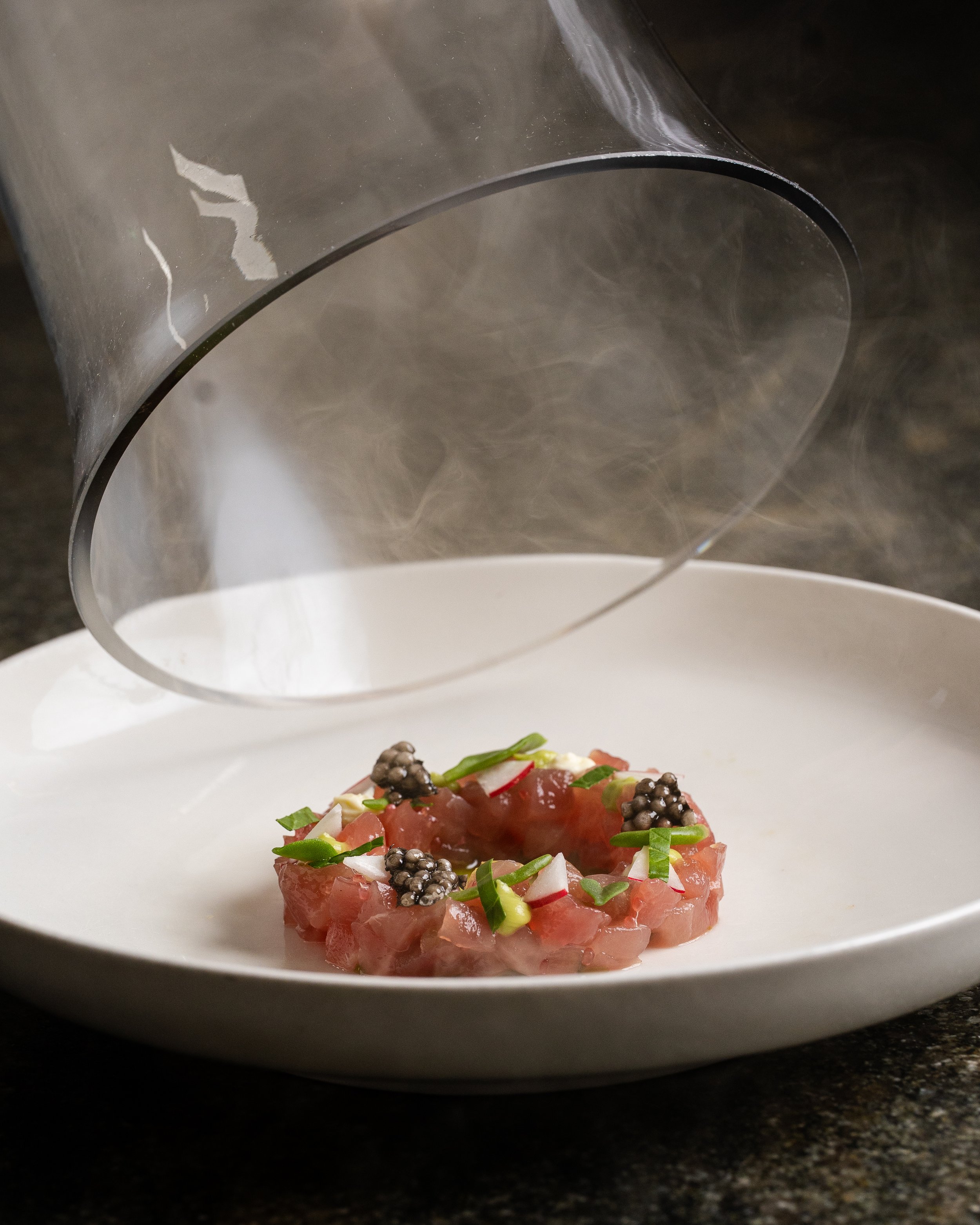 Tuna tartare garnished with chopped green onions, radish slices, and black caviar, with a glass dome overhead.