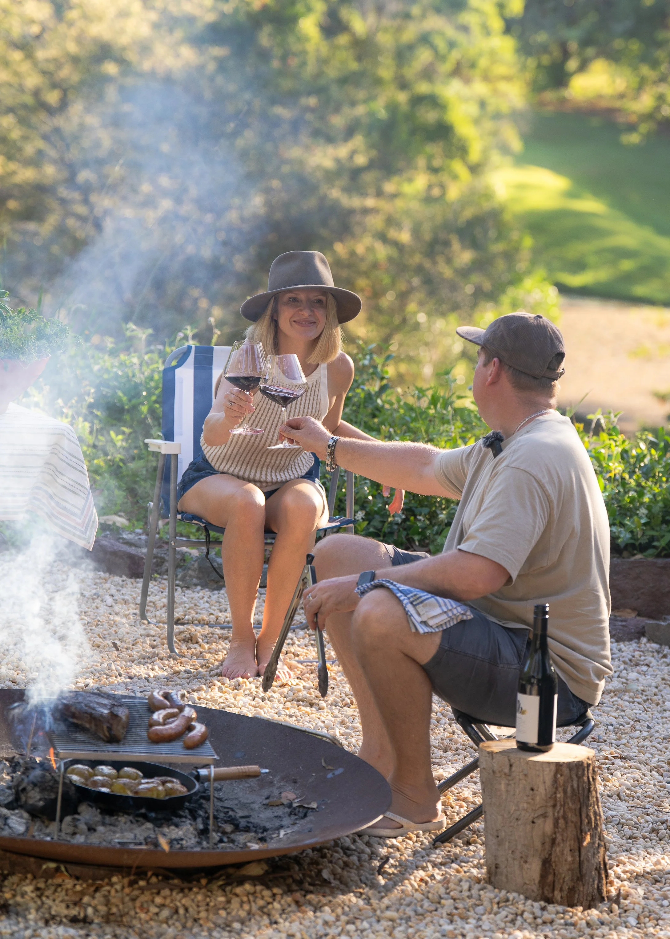 A couple enjoying a backyard barbecue and wine on a sunny day, with the woman sitting in a foldable chair, clinking glasses with the man who is seated on a small stool, with a grill and sausage in the foreground.