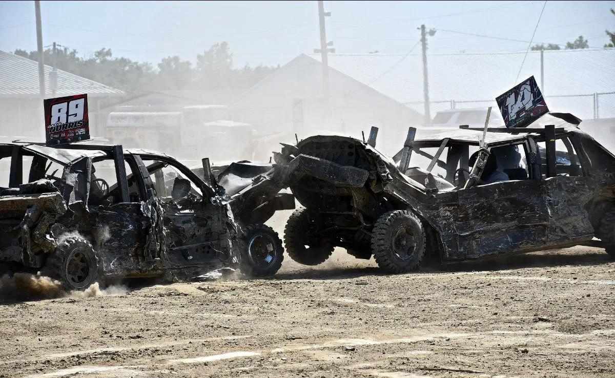 Two severely damaged race cars collide on a dirt track during a race. Dust and debris are flying, with race numbers visible on the cars.