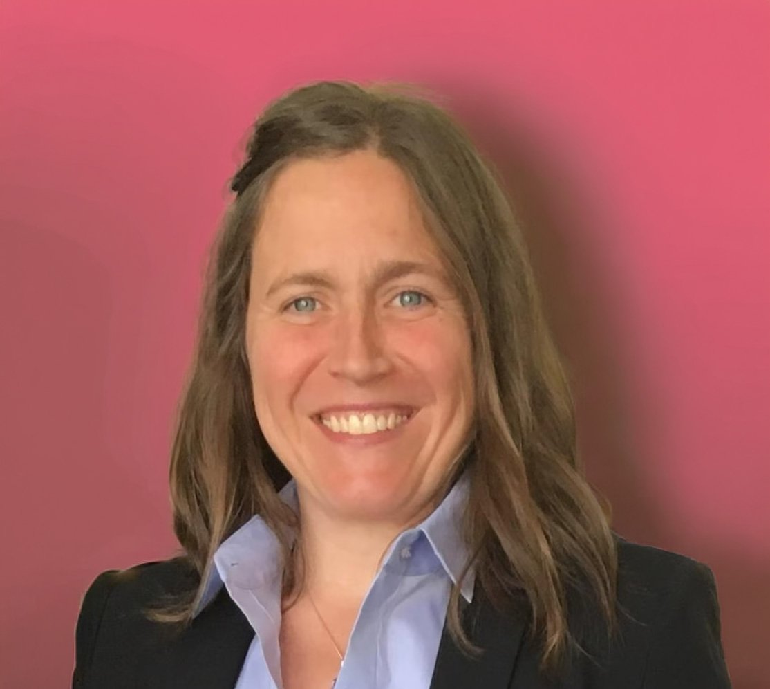 Stephanie Engelsman. Professional woman with shoulder-length brown hair, blue eyes, wearing a black blazer and light blue shirt, smiling against a pink background.