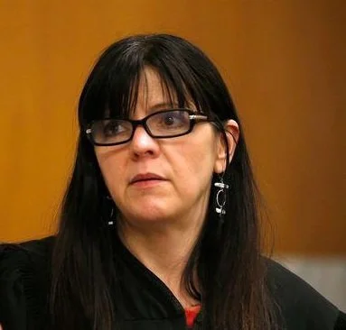 Judge Cheryl Albrecht. A woman with long dark hair, glasses, and earrings looking to the side