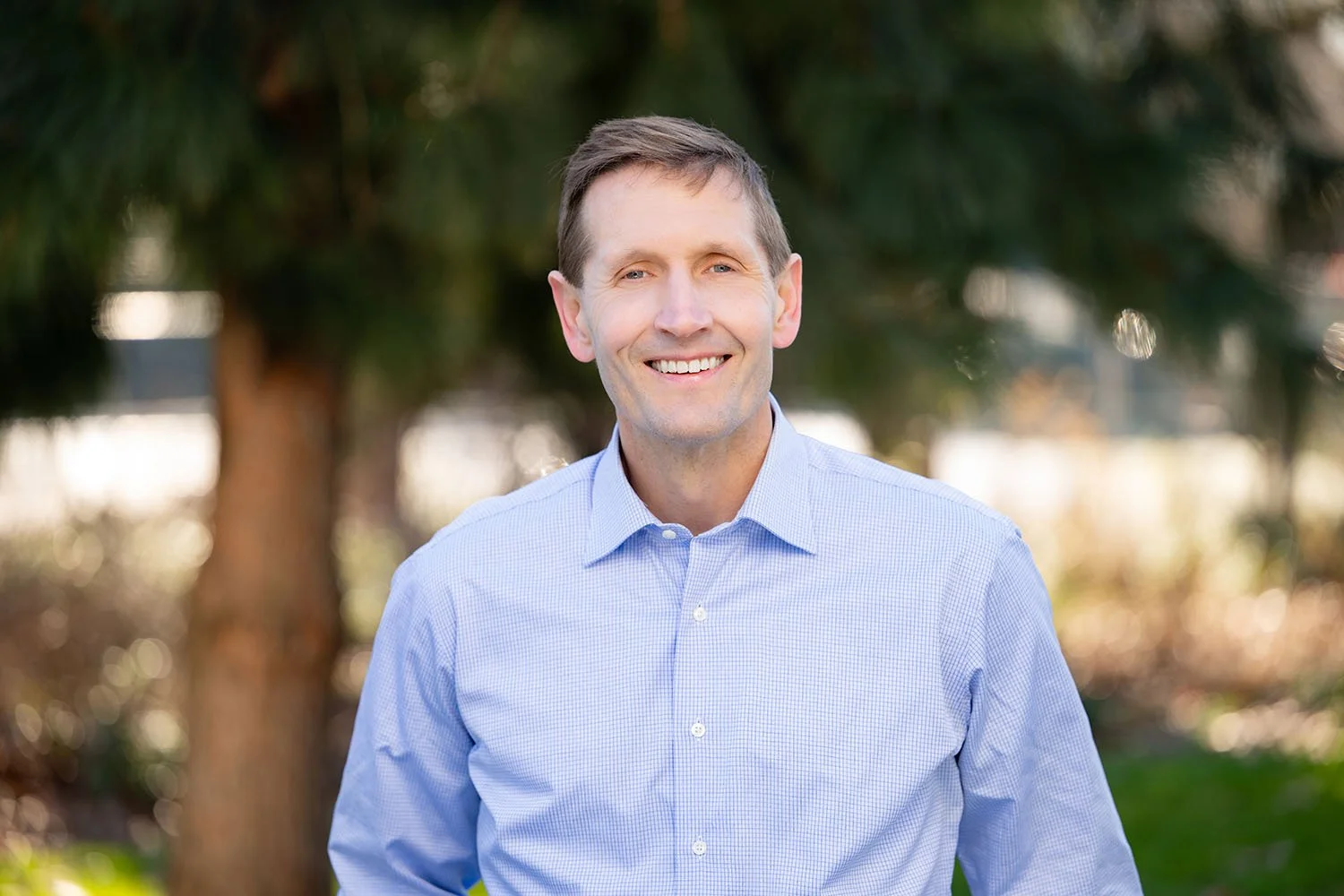 A smiling man with short brown hair wearing a light blue dress shirt standing outdoors in front of trees.