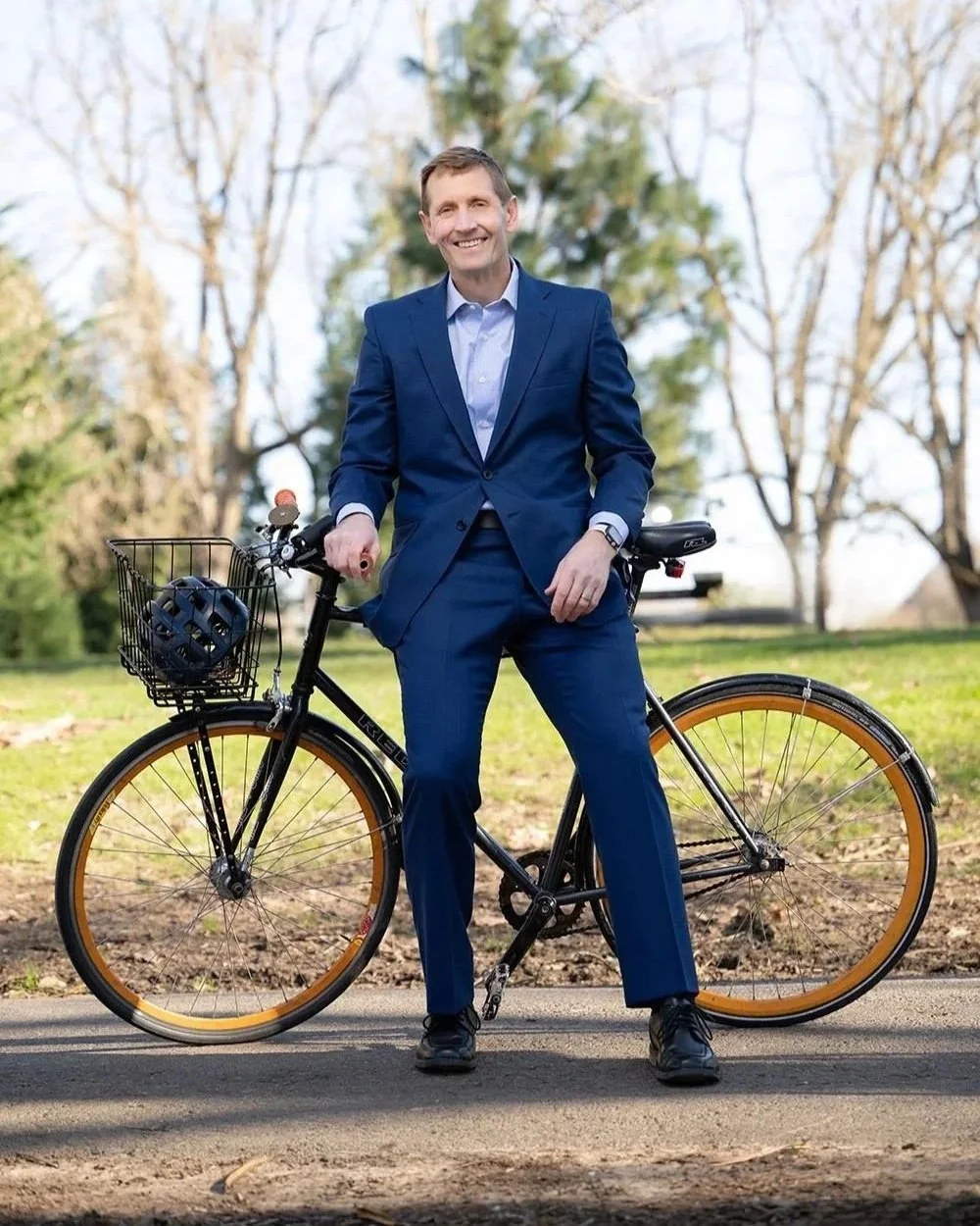 A man in a blue suit standing outdoors next to a black bicycle with a front basket, smiling. The background features leafless trees and a partly cloudy sky.