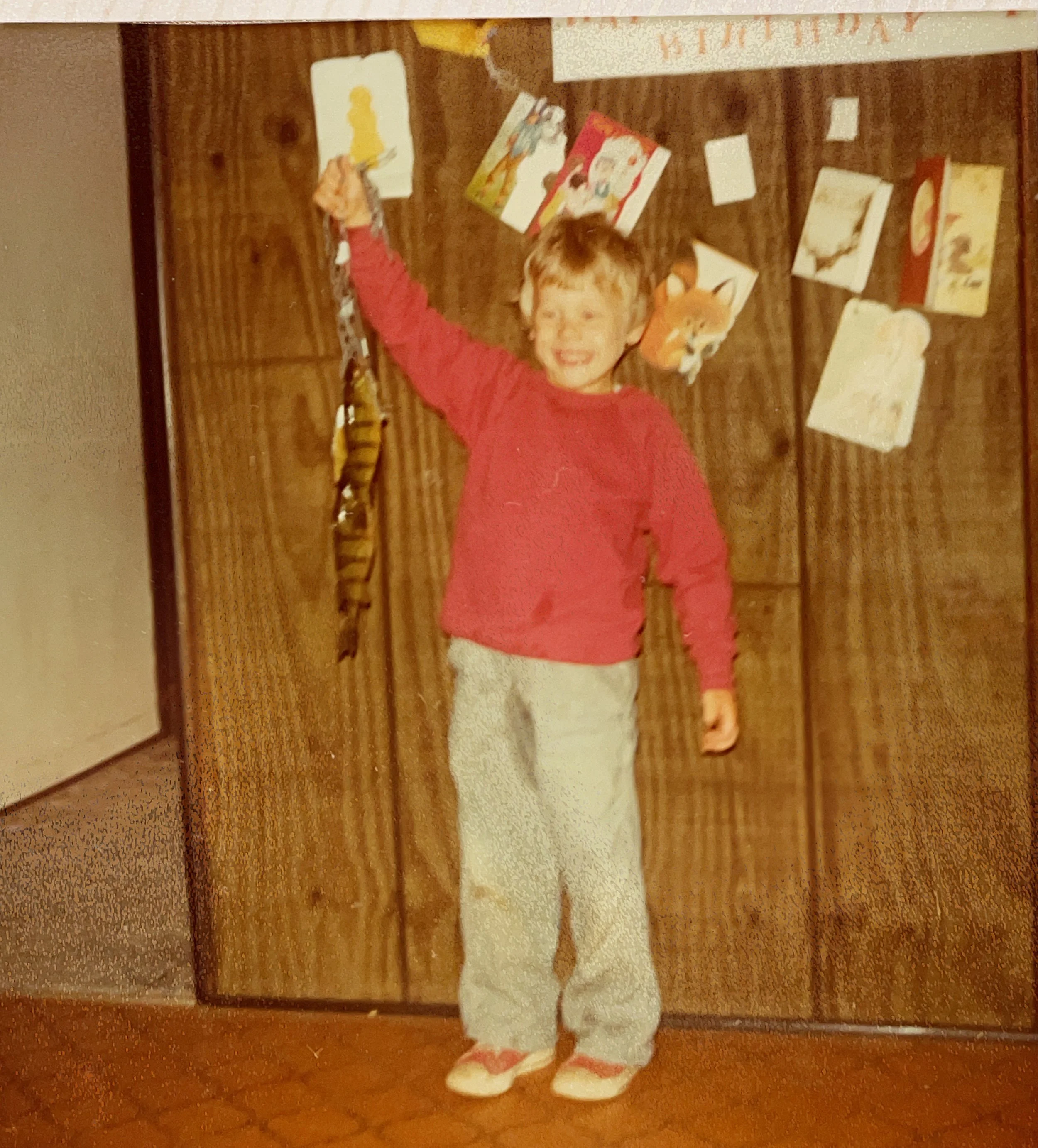A young boy with red hair and a red sweater smiling and holding a string of fish in front of a wooden door decorated for a birthday with birthday cards and a