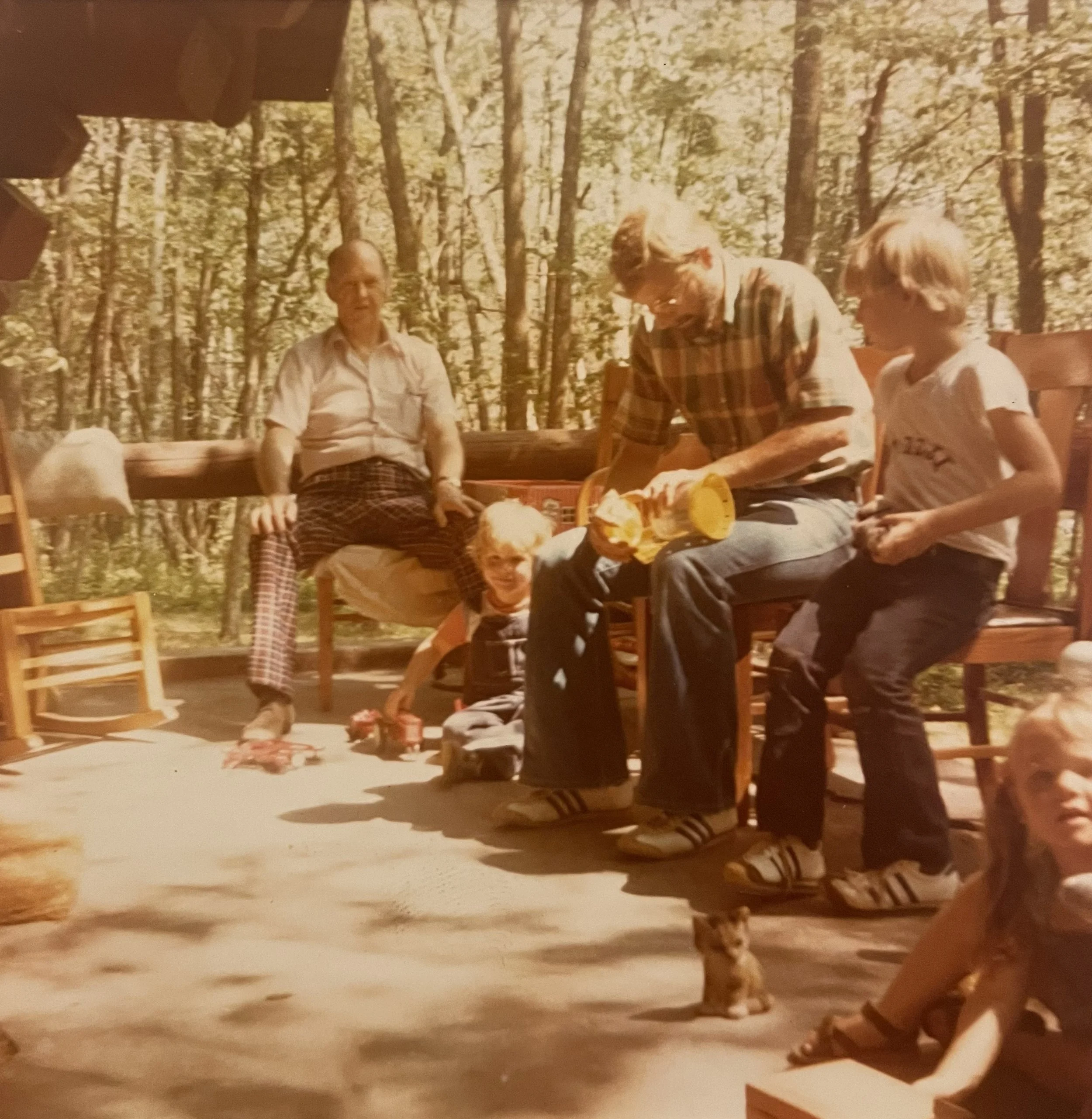 A vintage photo of two men and three children sitting in a wooded outdoor setting. One man is seated on a bench, the other is unwrapping a gift and the children are playing with toys.