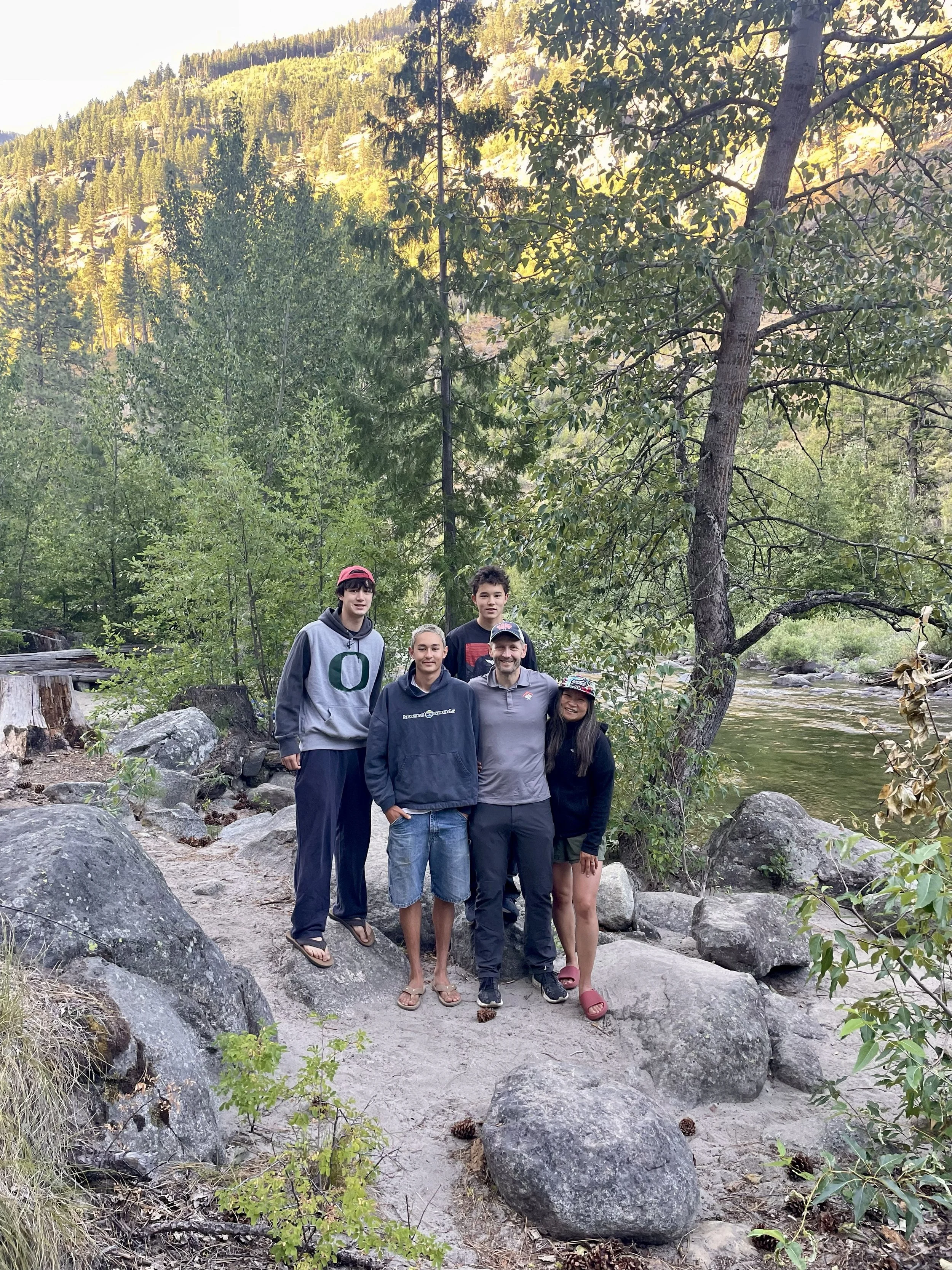 Group of five people standing on rocks near a river in a forested area with mountains in the background.