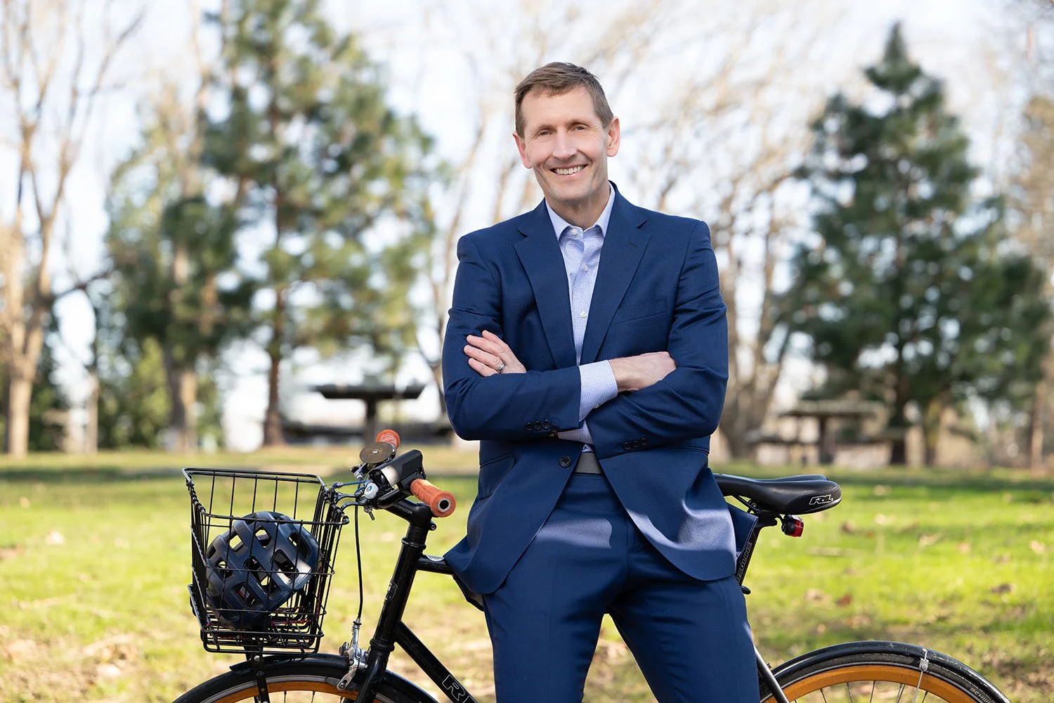 A man in a blue suit standing outdoors with crossed arms, smiling, next to a black bicycle with a helmet in the front basket, trees, and a park in the background.