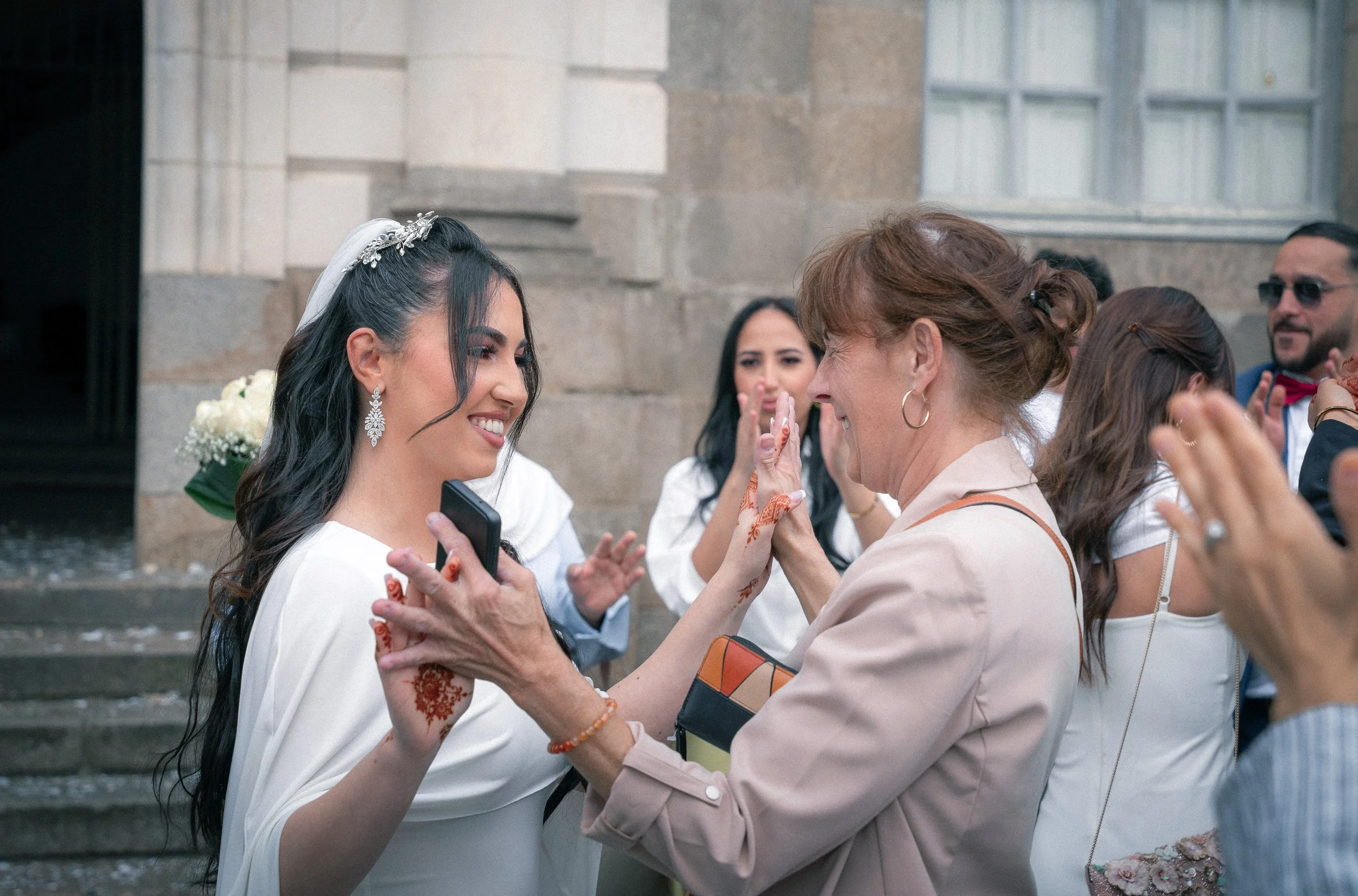 Une jeune femme en robe de mariée souriante échange un moment émouvant avec une femme plus âgée, lors d'une célébration en extérieur, entourée d'autres personnes qui semblent joyeuses.
