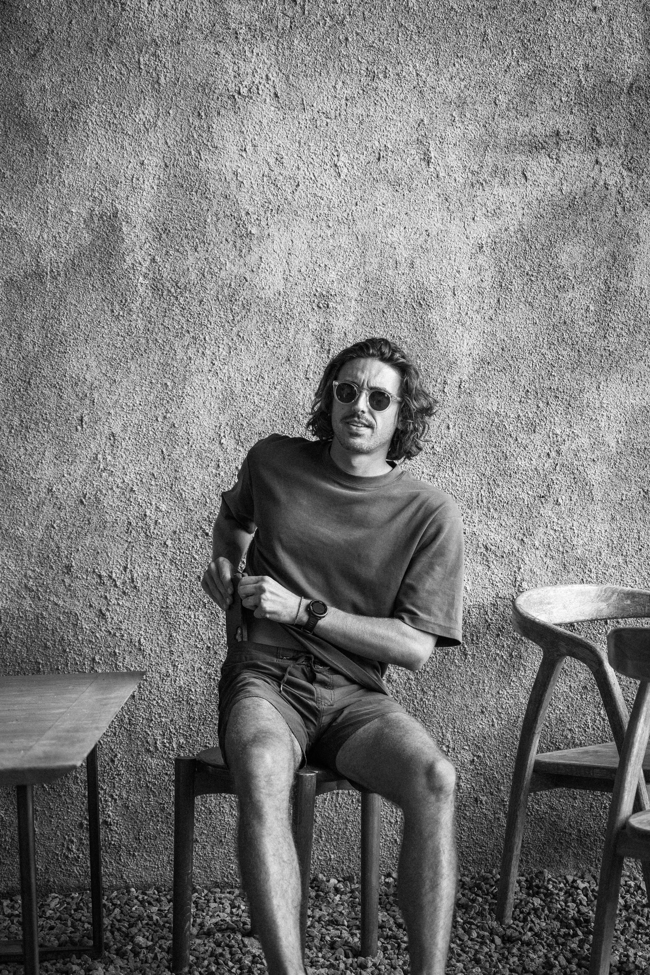 Un jeune homme aux cheveux longs, portant des lunettes de soleil, une chemise à manches courtes et un short en jean, est assis sur une chaise en bois devant un mur en béton. Il regarde vers l'appareil photo, avec un style décontracté.