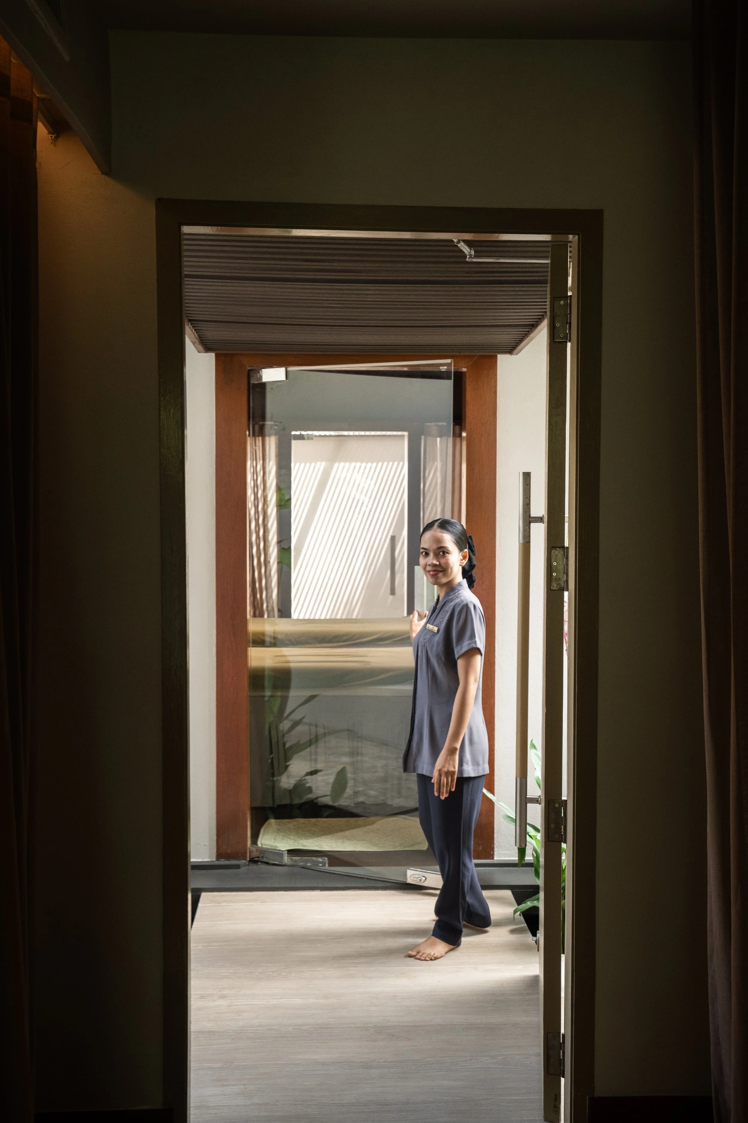 Une femme d'affaires souriante, habillée en uniforme professionnel, se tient à l'entrée d'une pièce, vue à travers une porte entrouverte, dans une ambiance intérieure avec un sol en bois clair et un décor minimaliste.