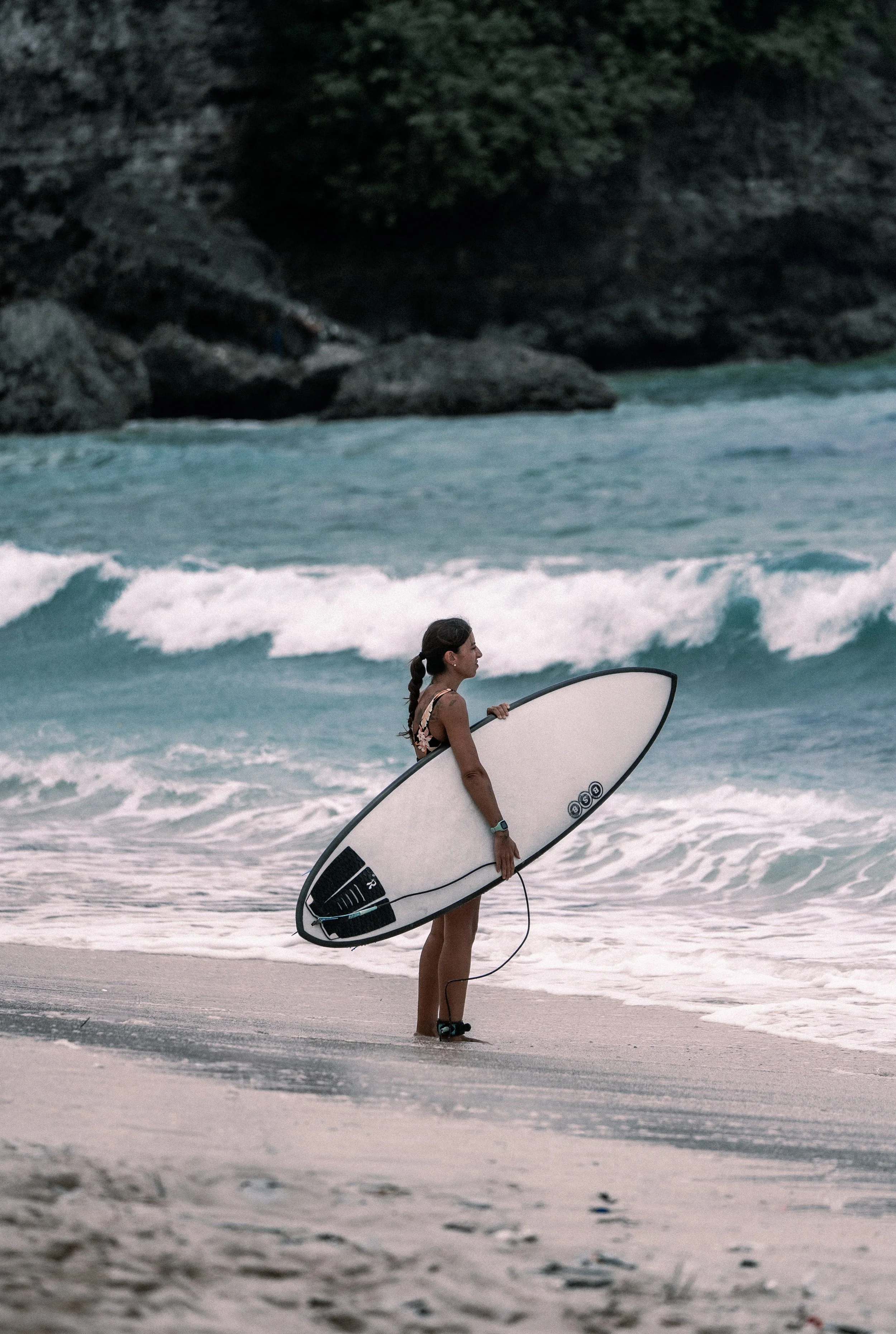 Jeune femme debout sur la plage, tenant une planche de surf, regardant l'océan.