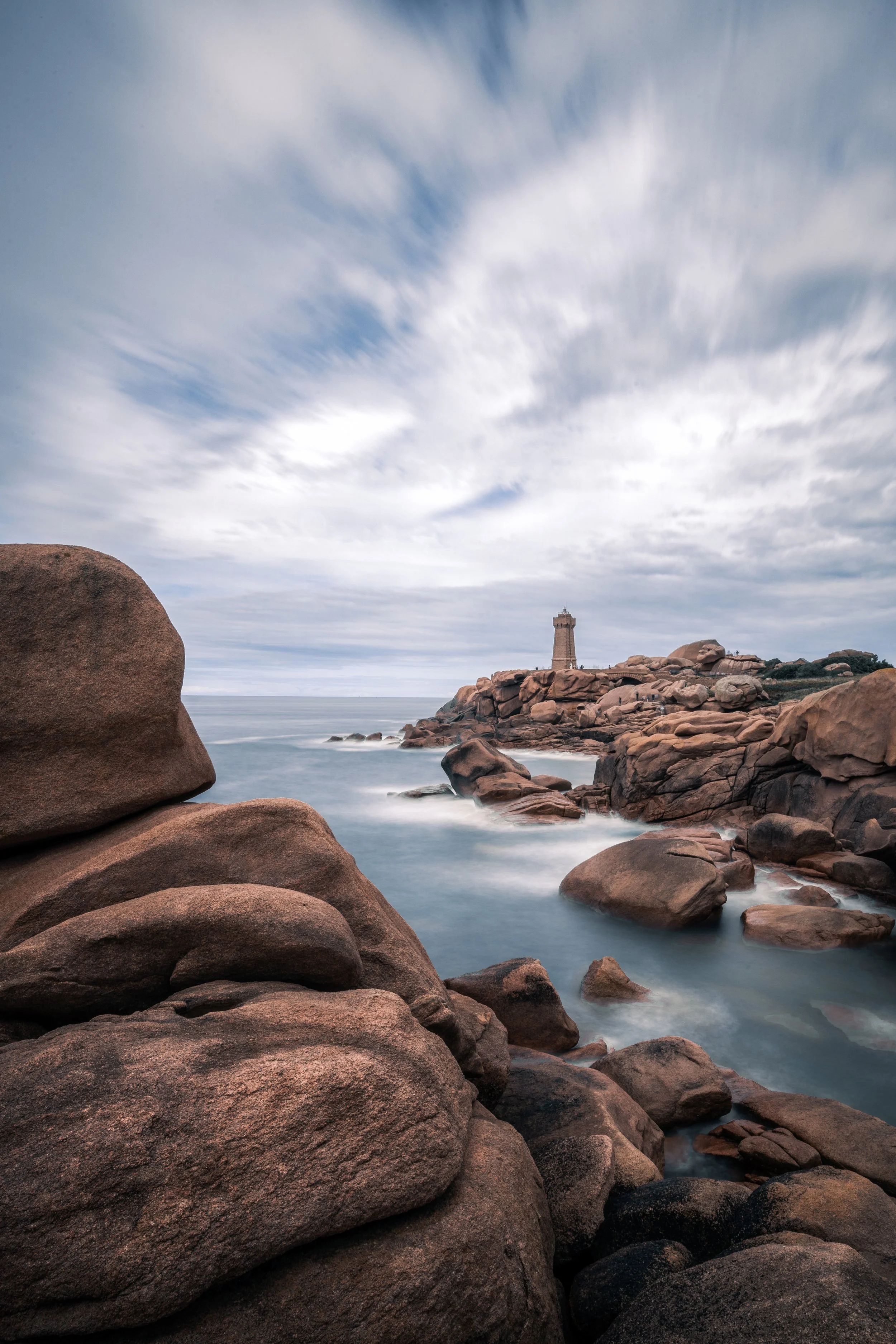 Côte rocheuse avec des gros rochers au premier plan, un phare en pierre sur la falaise, et la mer calme sous un ciel nuageux.