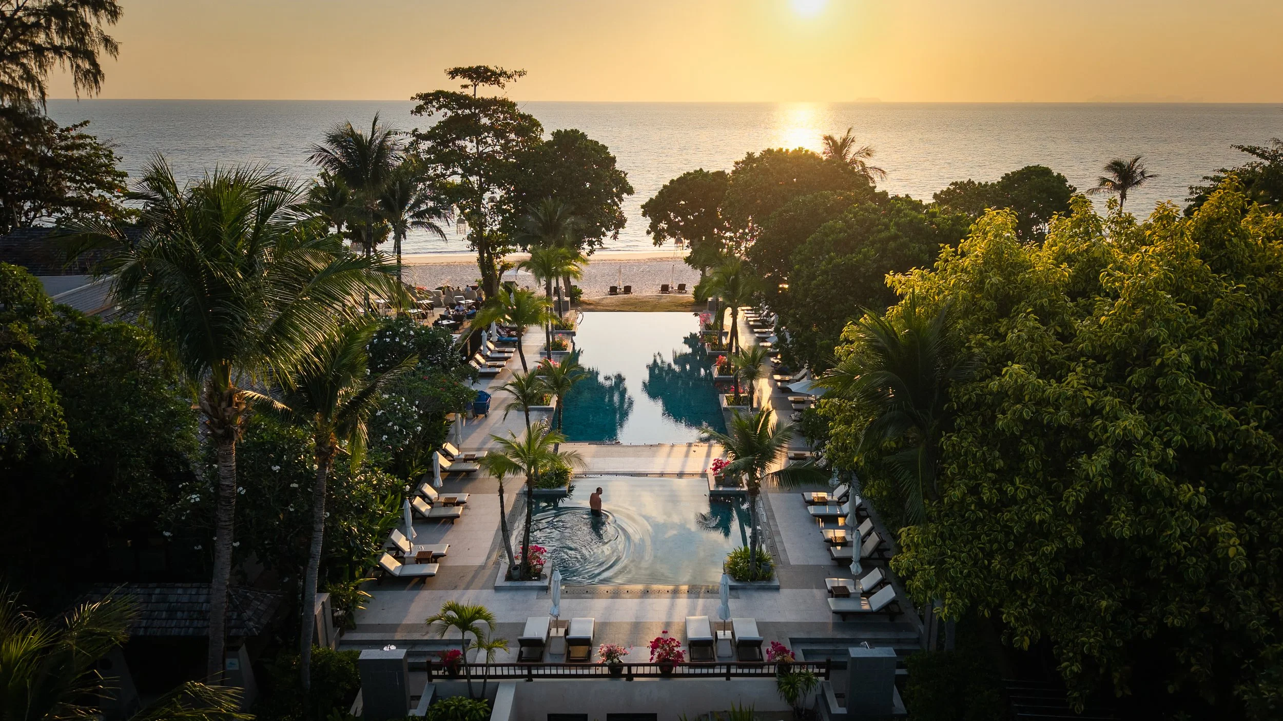 Piscine de luxe avec transats entourée de palmiers, en face de la mer au coucher du soleil.