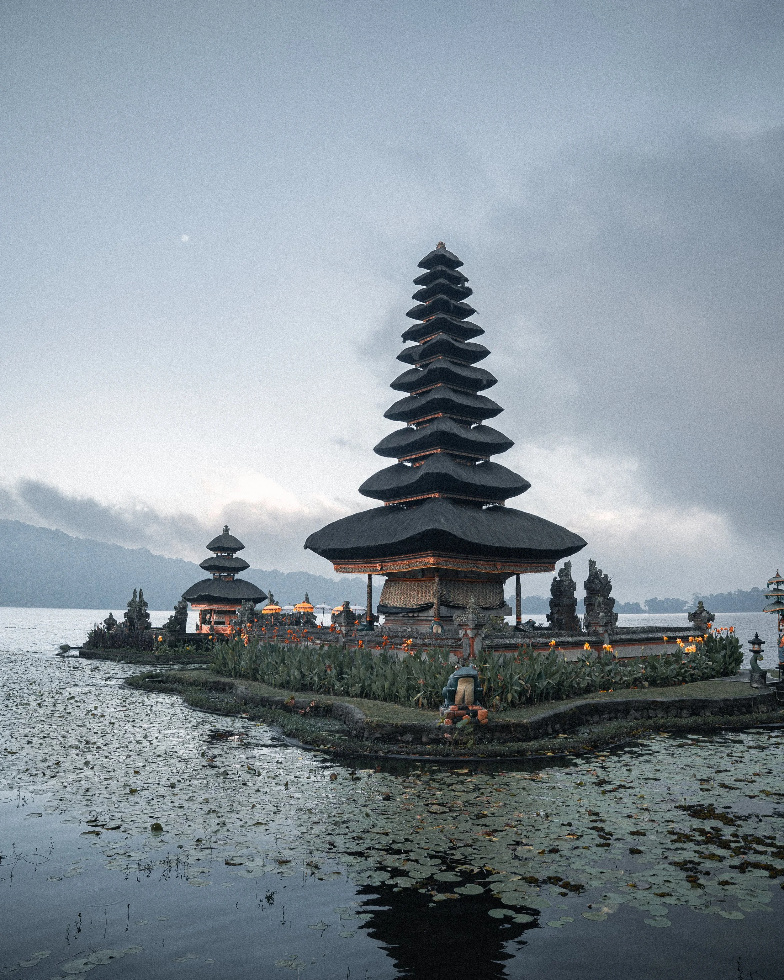 Une pagode traditionnelle en pierre avec plusieurs toitures en forme de pagode, située sur une petite île dans un lac avec des nénuphars flottants, au coucher du soleil ou en début de soirée.